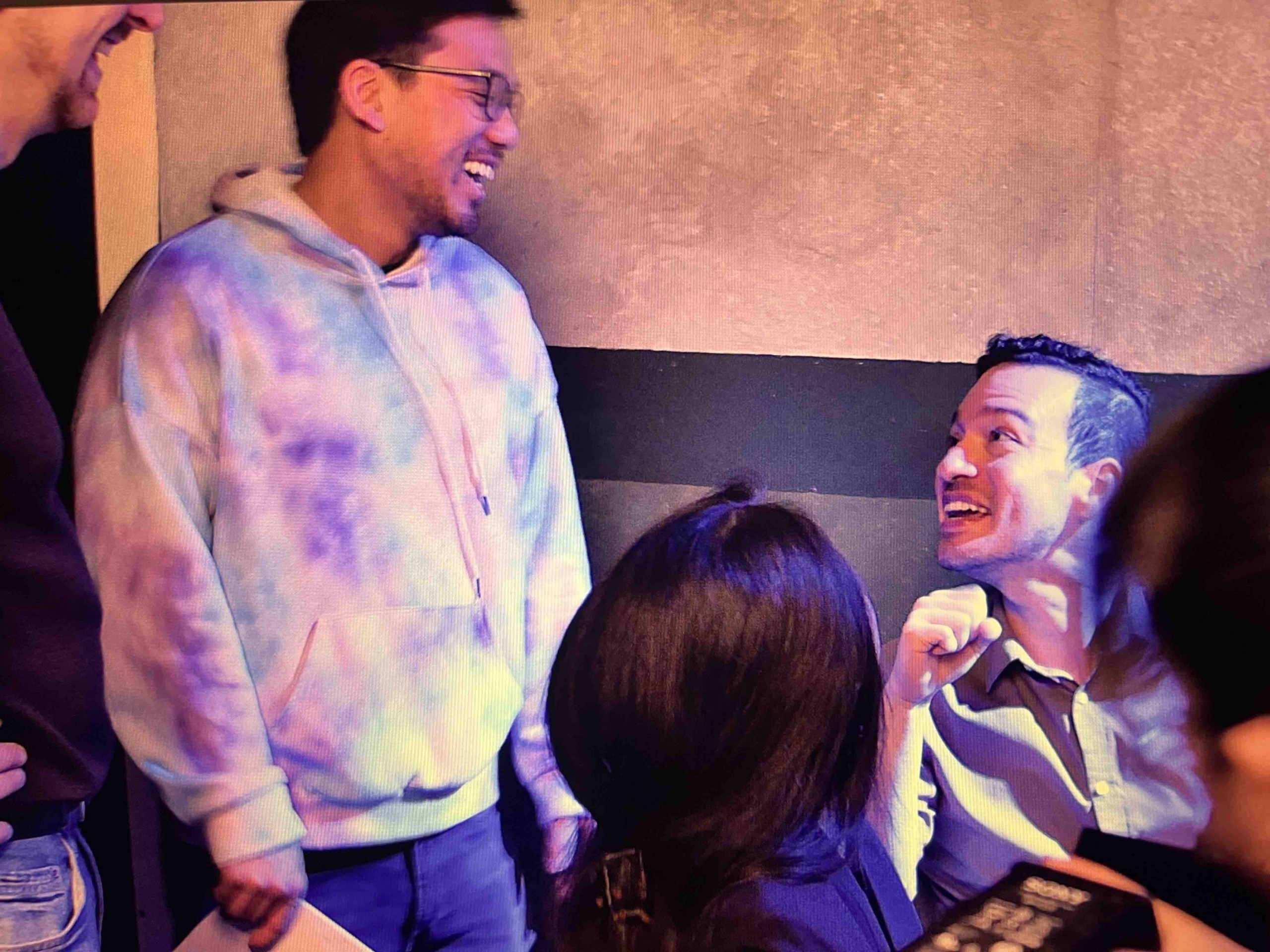 Two men are smiling and talking, one wearing a tie-dye hoodie and the other in a striped shirt, with a woman in the foreground.