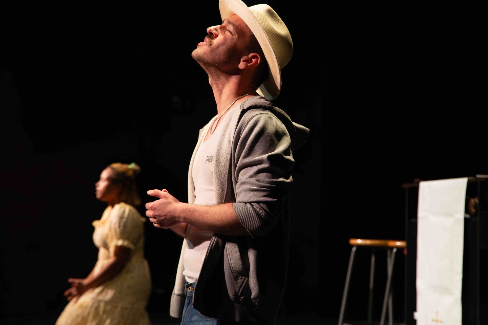 Man wearing a hat and hoodie, standing on stage with eyes closed, in a dark setting, with a woman in the background.