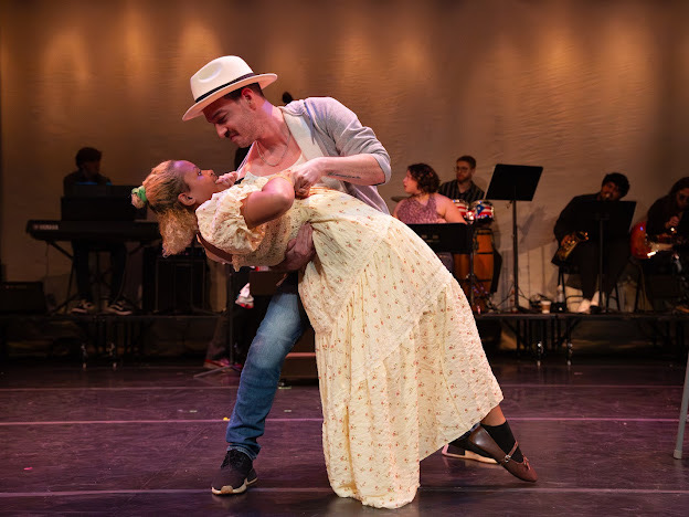 A man and woman dance on stage with musicians in the background, the man wearing a hat and the woman in a long dress.