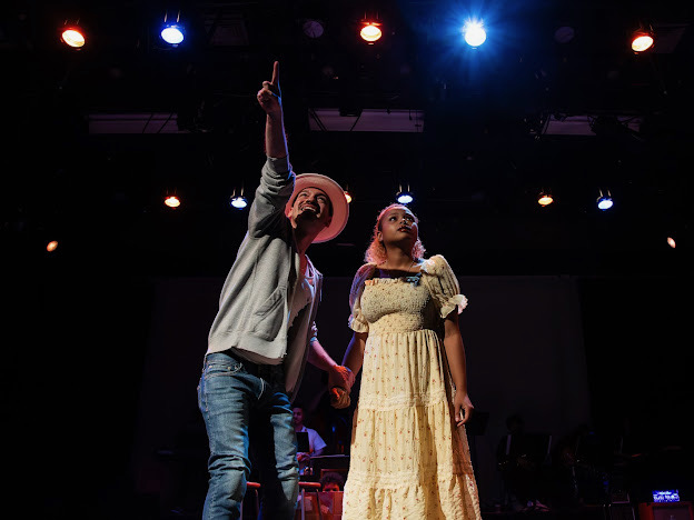 Two performers on stage, one with a hat pointing upward, the other in a yellow dress, stage lights above, dark background.