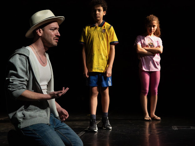 Man with a hat and gray hoodie sitting on stage, two children standing with arms crossed, dark background