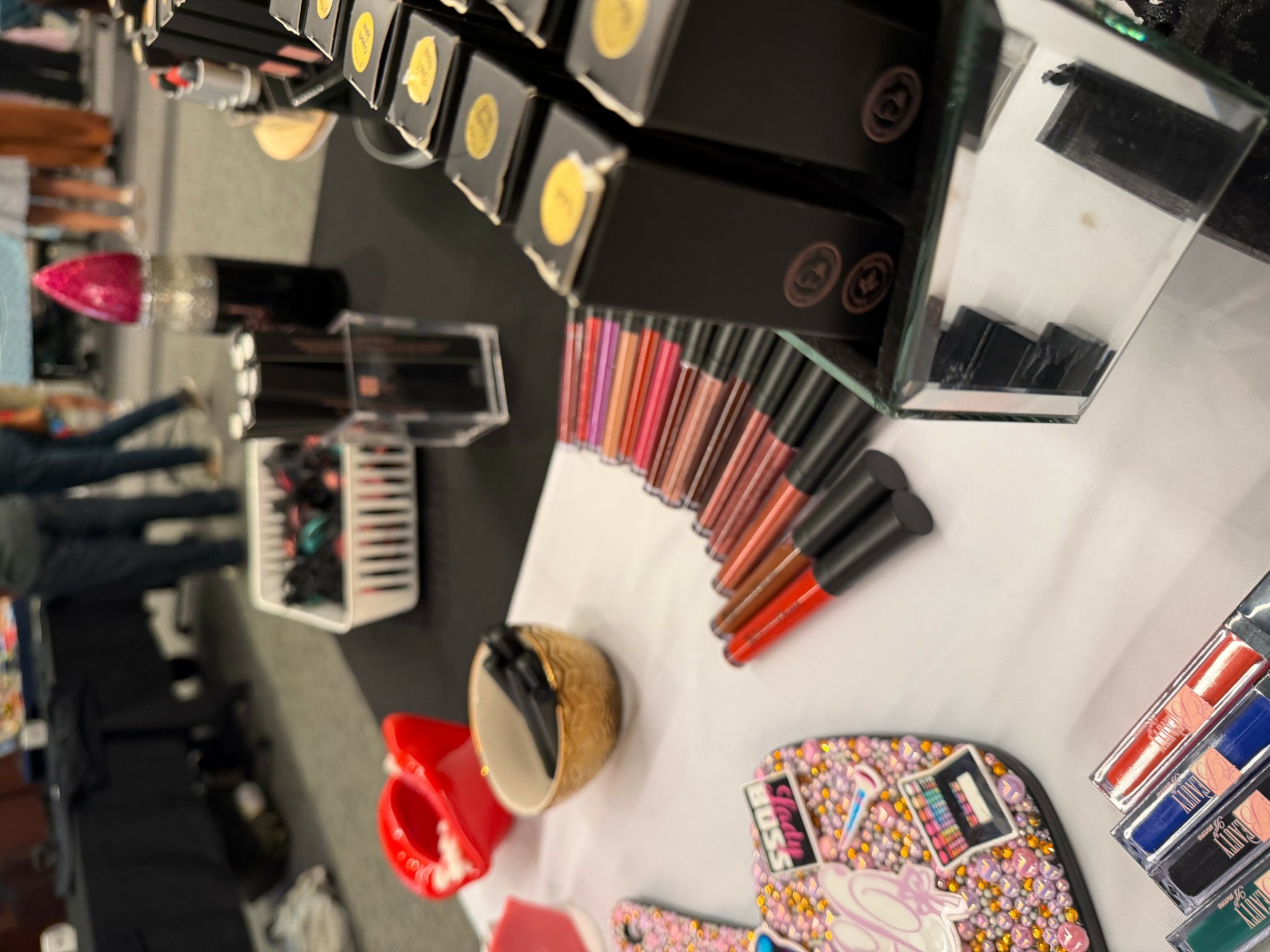 Lipsticks and makeup products arranged on a white surface with people in the background.
