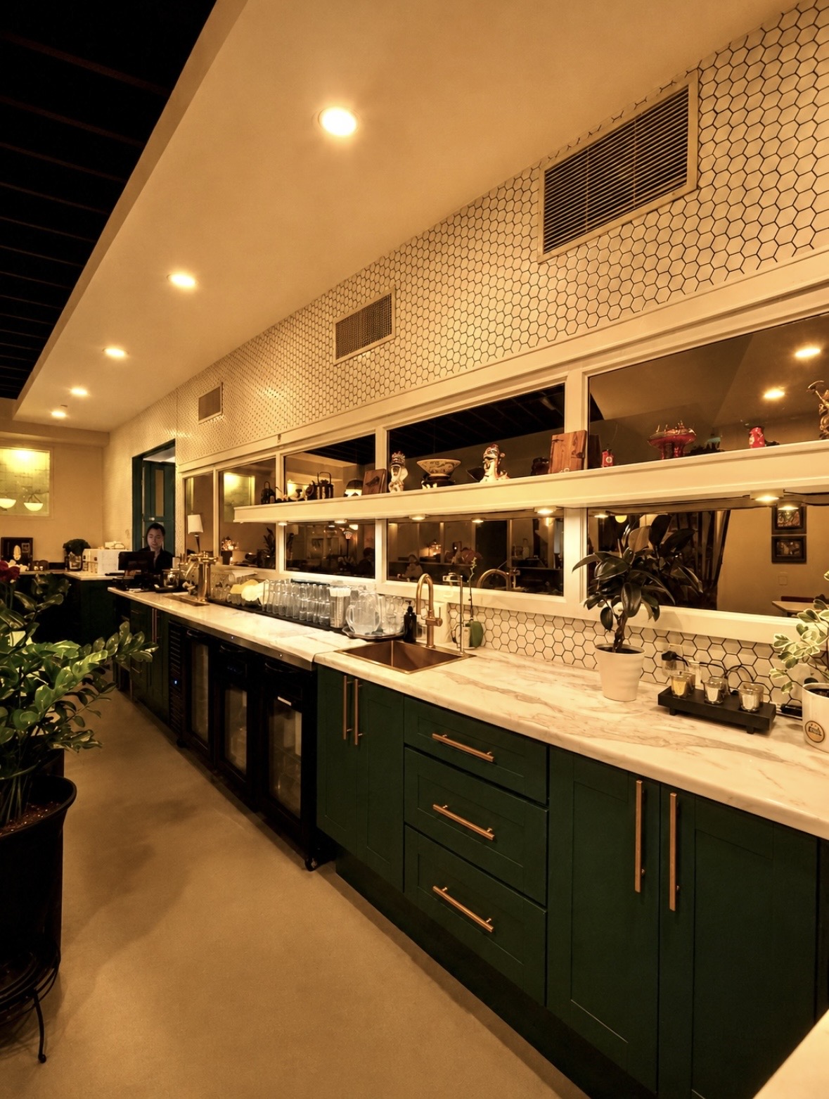 Kitchen with green cabinets, white countertop, and large windows, illuminated by ceiling lights, with plants and kitchen items visible.