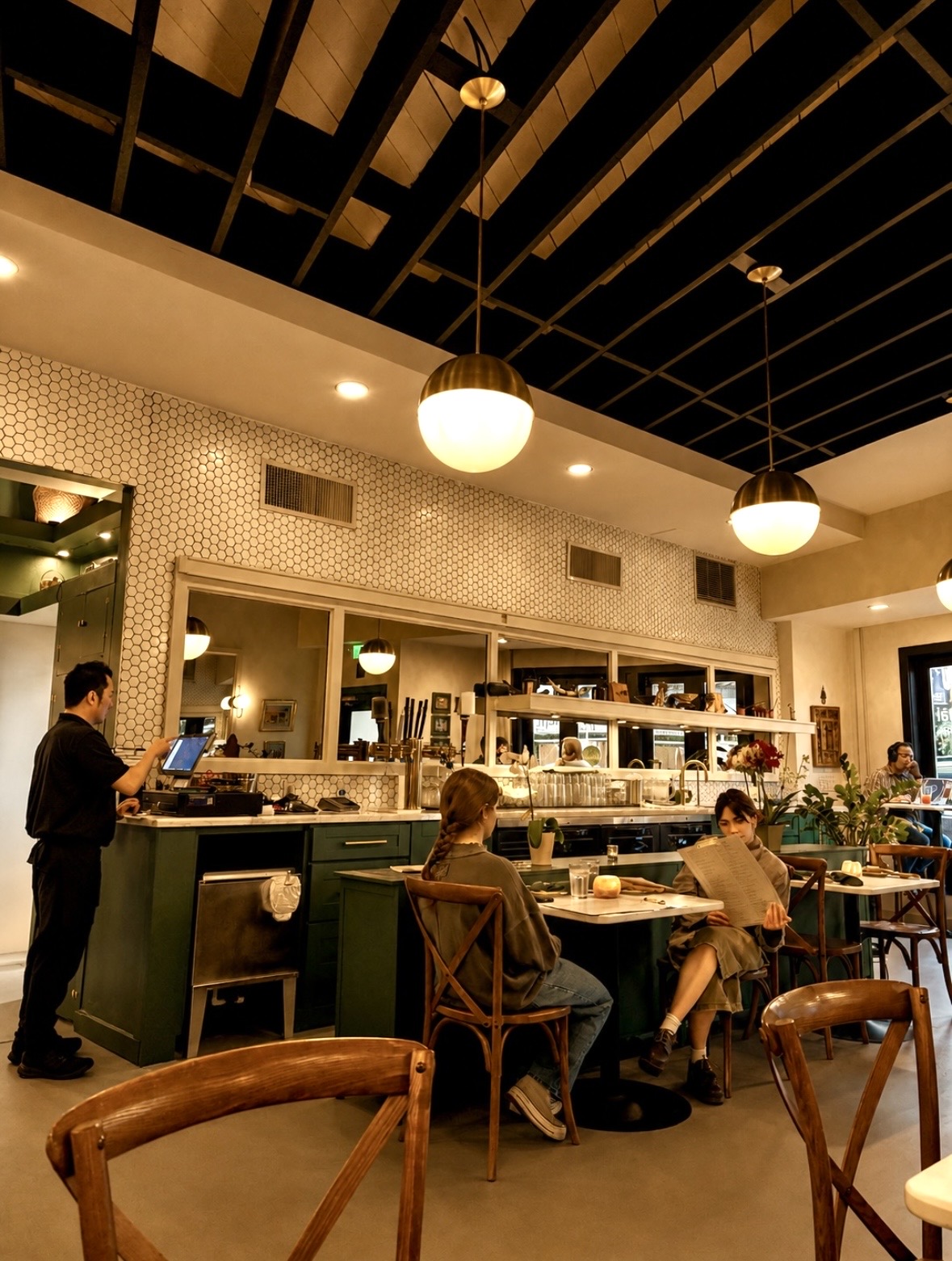 People sit at a bar counter in a cafe with hanging lights and a black ceiling, while a staff member works behind the counter.