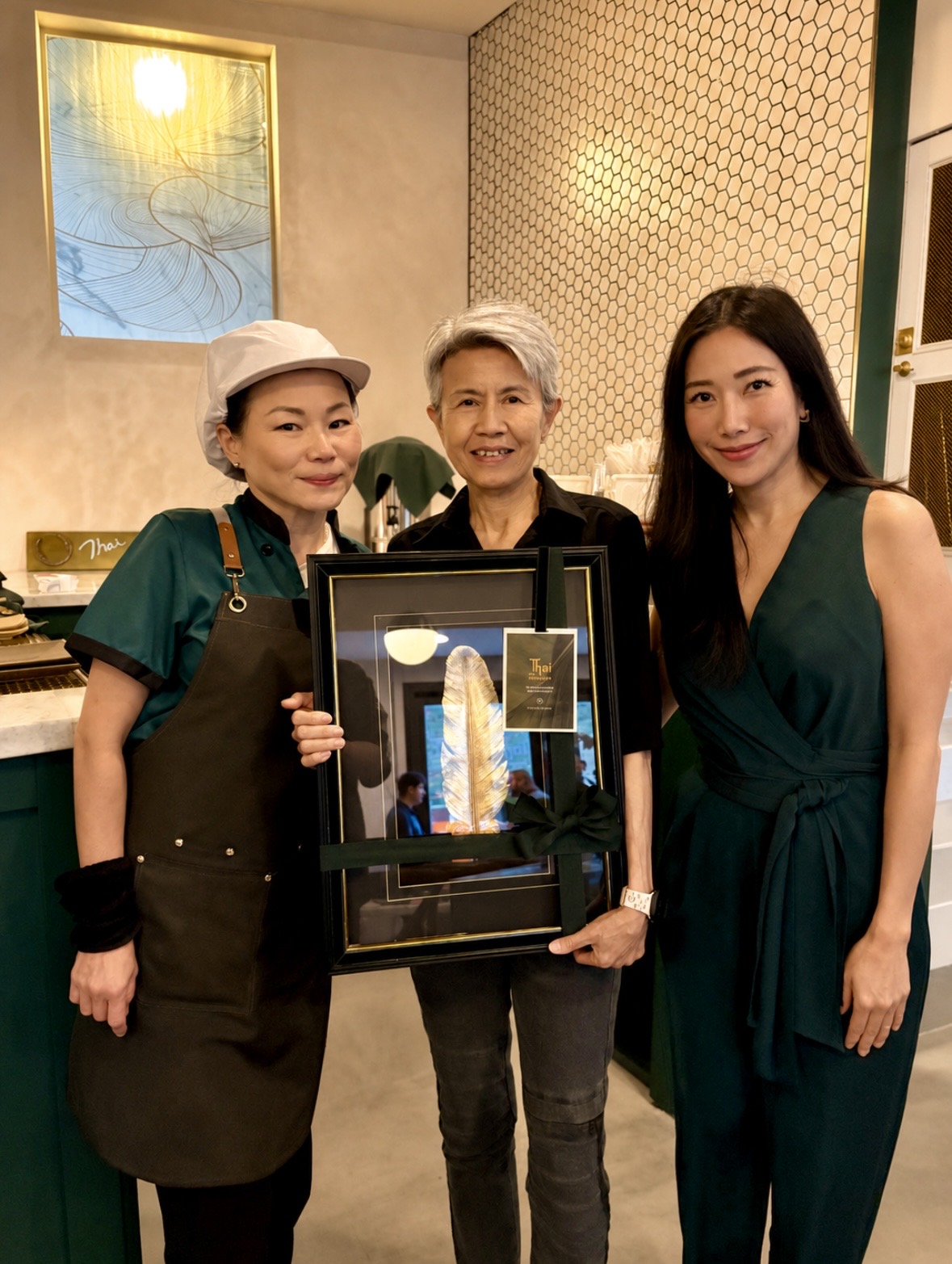 Three women standing indoors, one holding a framed award or artwork, smiling at the camera. The background includes a window and patterned wall.