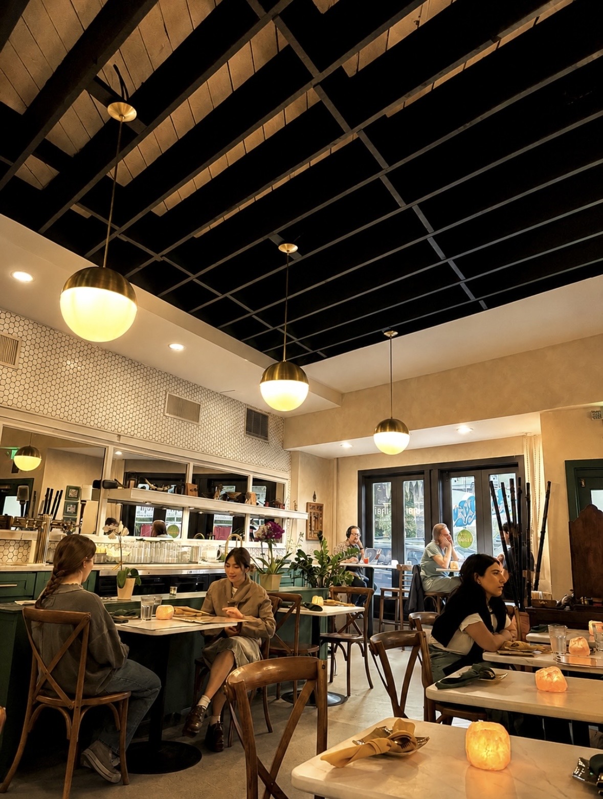 People dining in a restaurant with black ceiling, hanging lights, and large windows, some engaged in conversation, candles on tables.