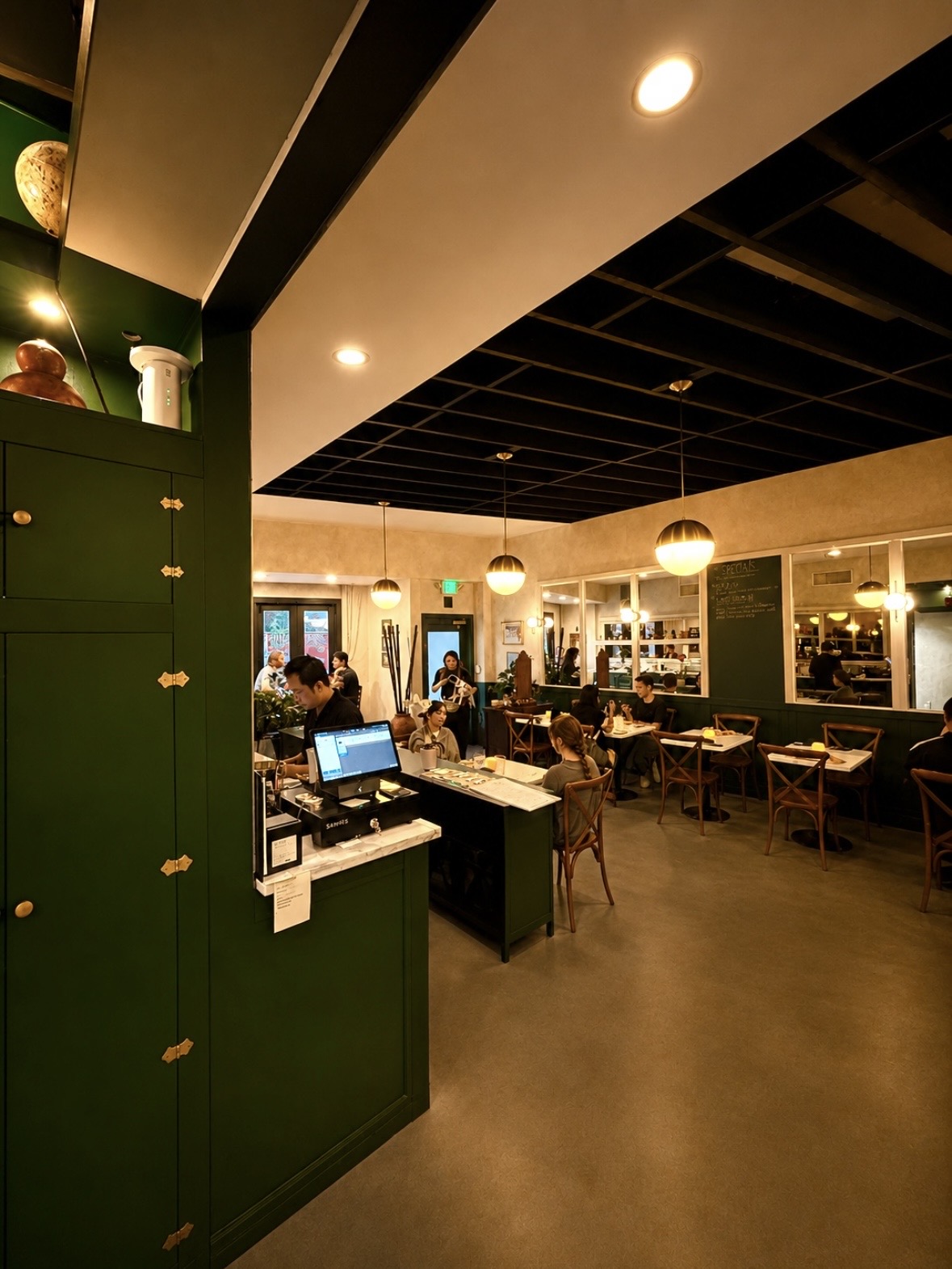 Interior of a cafe with people sitting at tables, green cabinets, and large windows, illuminated by ceiling lights.