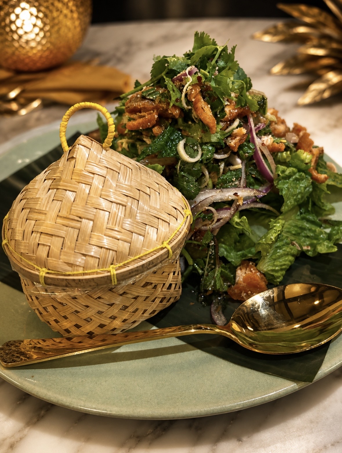 Salad with greens, vegetables, and toppings on a plate, with a woven container and a gold spoon nearby.
