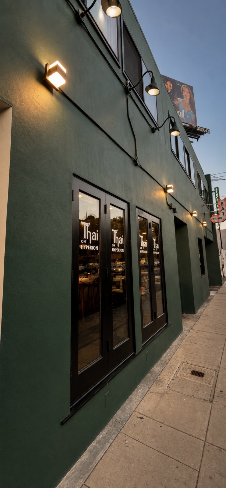 Green building exterior with large glass doors, outdoor lights, and a sidewalk, under a clear sky.