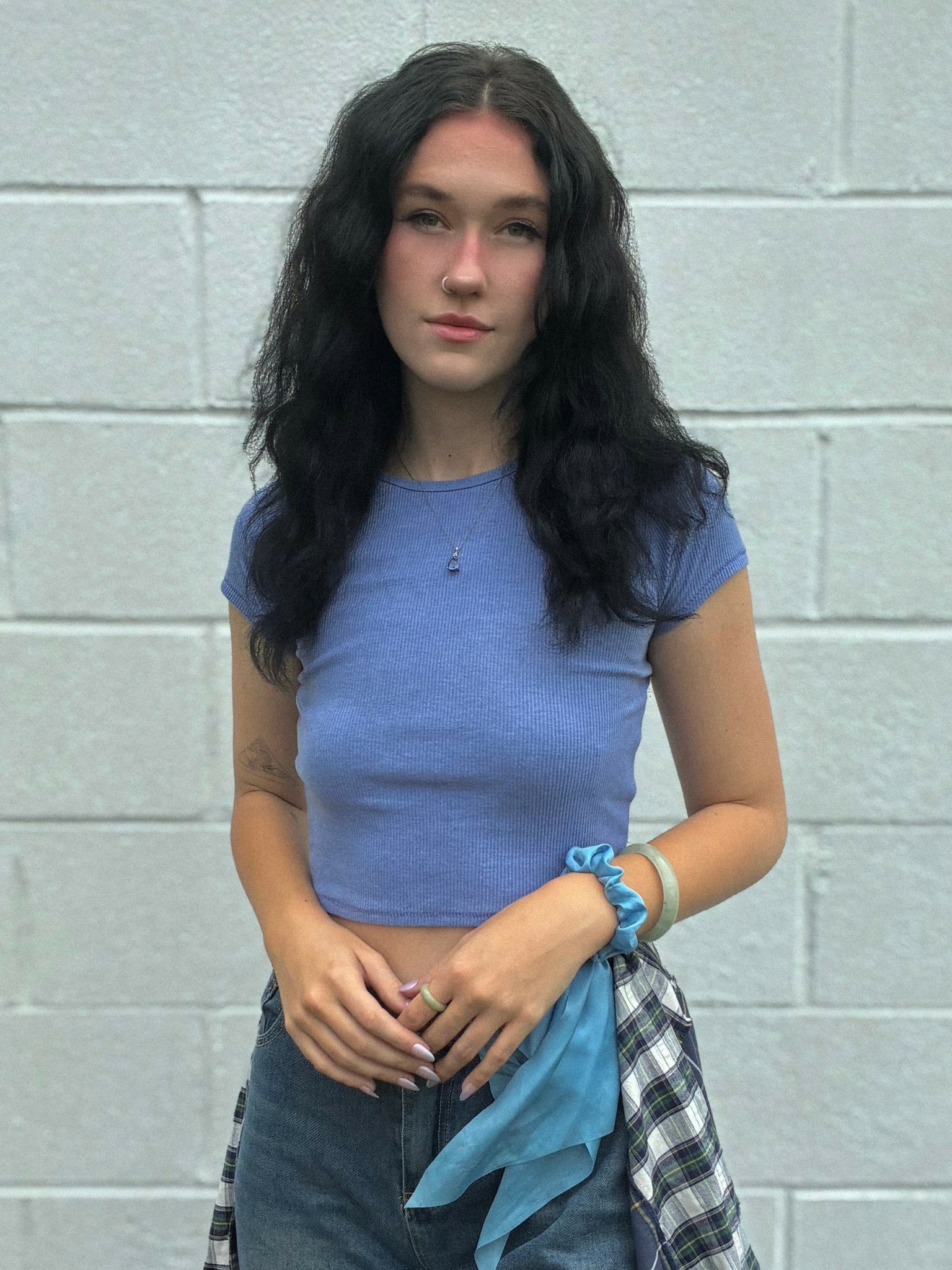 Young woman with long dark wavy hair wearing a blue crop top and jeans, standing against a light gray wall.
