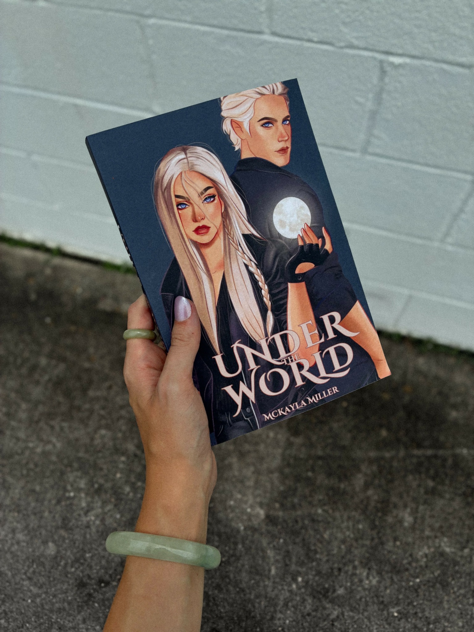 Hand holding a book titled 'Under the World' with two illustrated women on the cover, one with long blonde hair and the other with short white hair, against a light-colored wall.