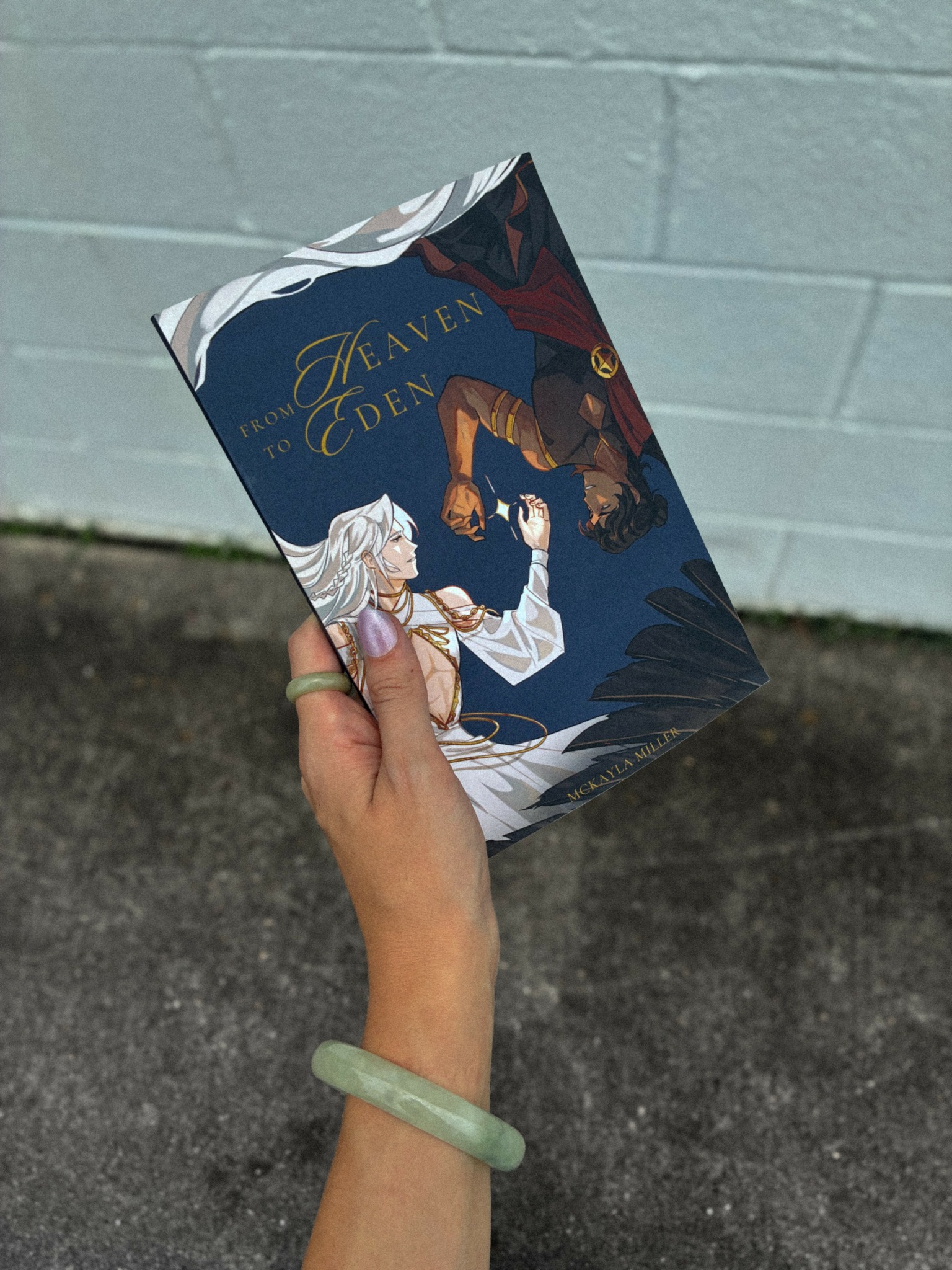Person holding a book with a blue cover and illustrated characters, standing outdoors against a light blue wall.