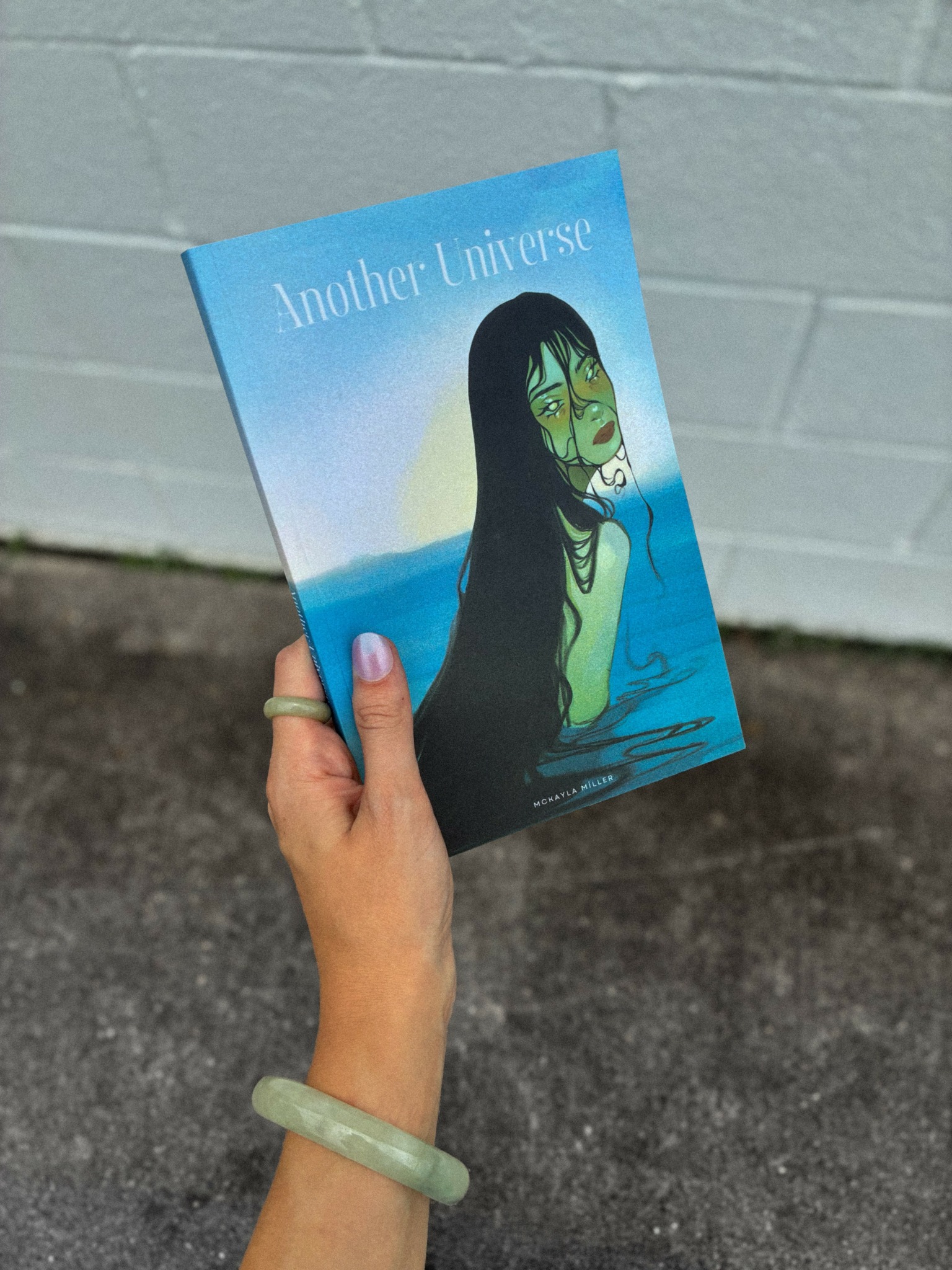 Person holding a book titled 'Another Universe' with an illustration of a woman with long black hair and green face paint, standing in water.