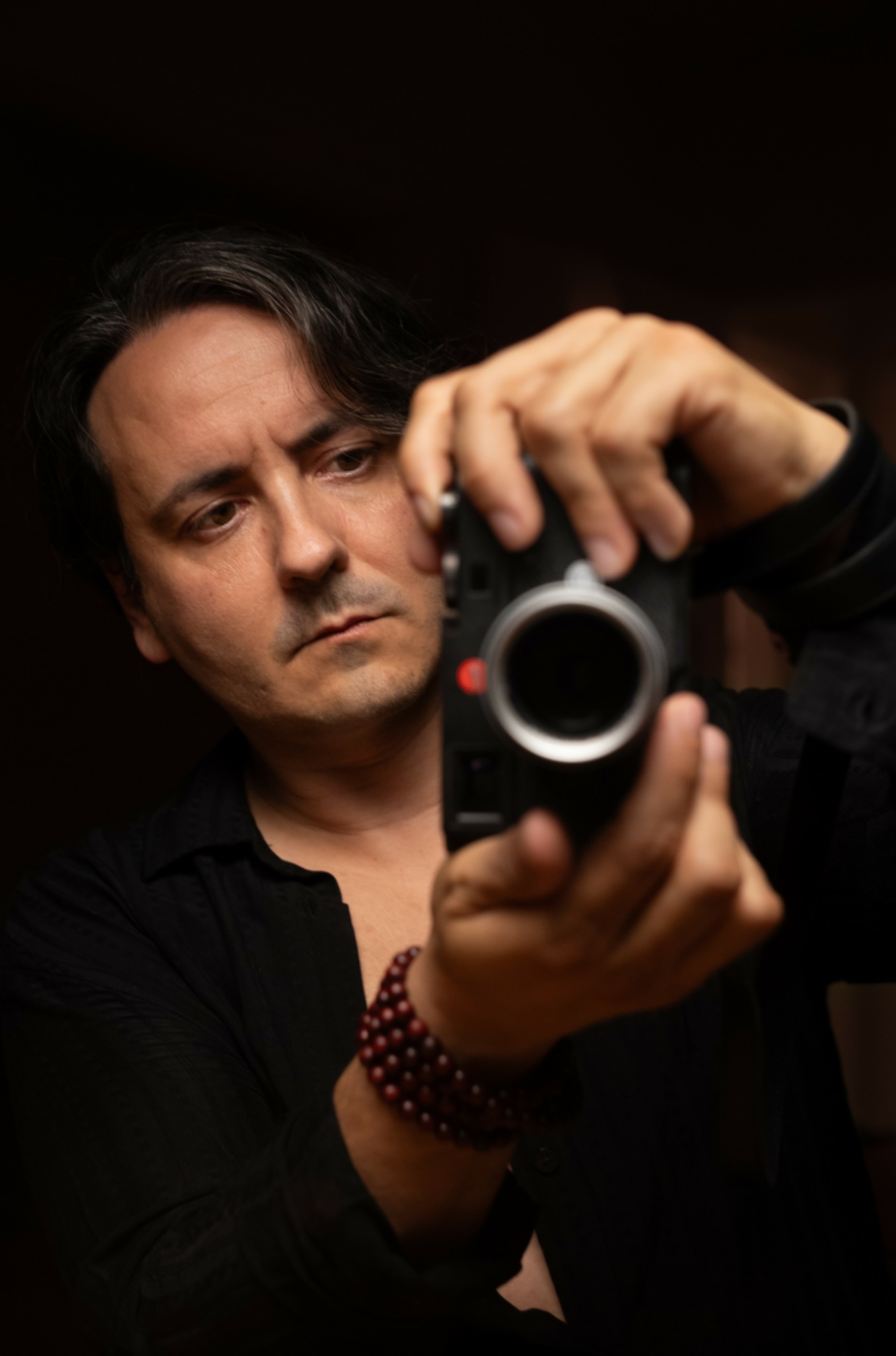 Man with dark hair taking a selfie with a camera, wearing a black shirt and a beaded bracelet, in a dark setting.