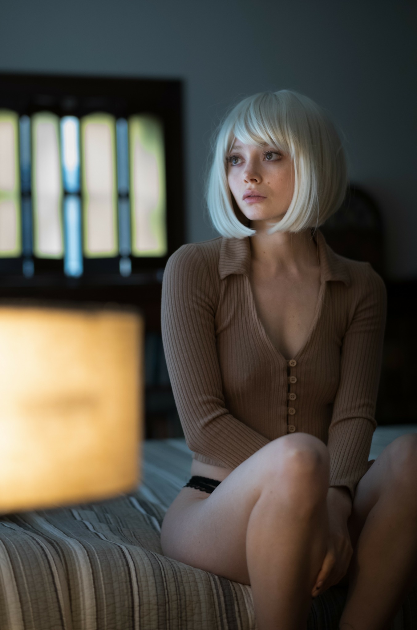 Young woman with short platinum blonde hair sitting on a bed in a dimly lit room, looking to the side.