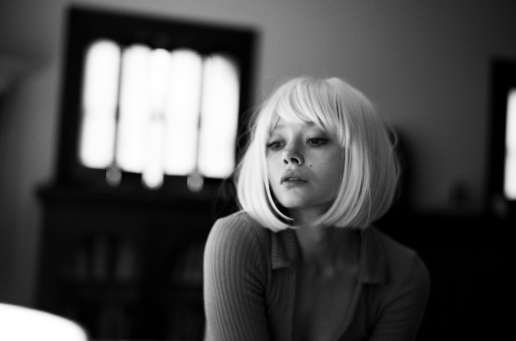 A young woman with a blonde bob hairstyle looks to the side in a dimly lit room.
