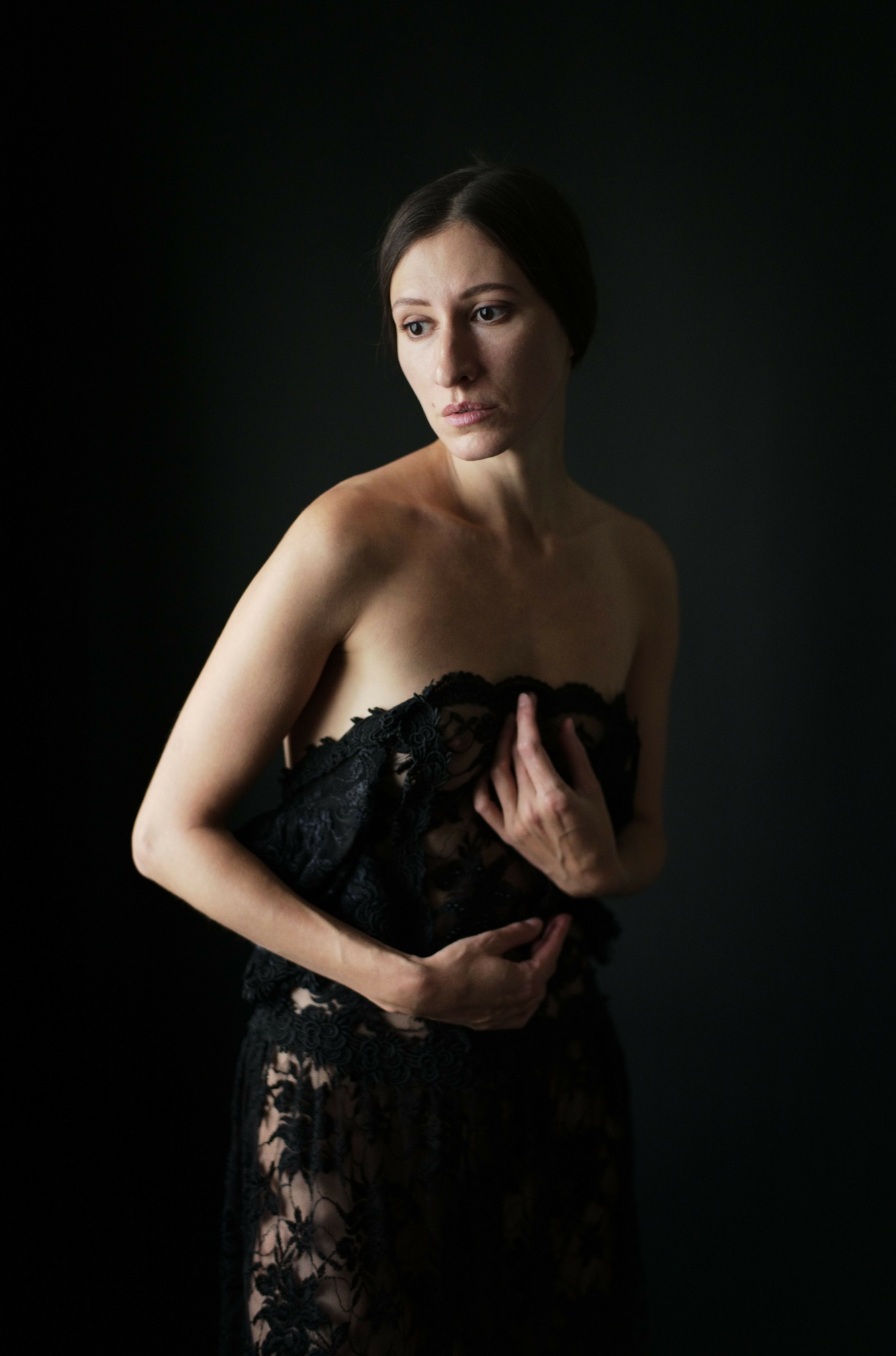 Woman in a black lace dress lying sideways against a dark background, looking at the camera with a neutral expression.