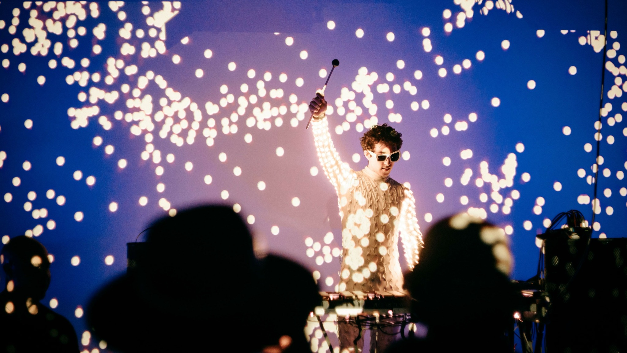 Performer on stage with a sparkling background, raising one arm, audience silhouettes in foreground.