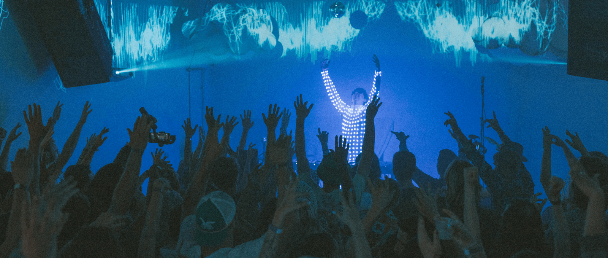 Person on stage with arms raised in front of an audience with hands raised, in a concert setting.