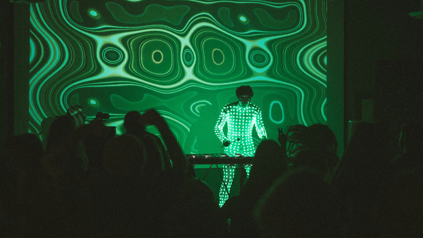 Person performing music on stage with a digital backdrop, audience watching in a dark room.