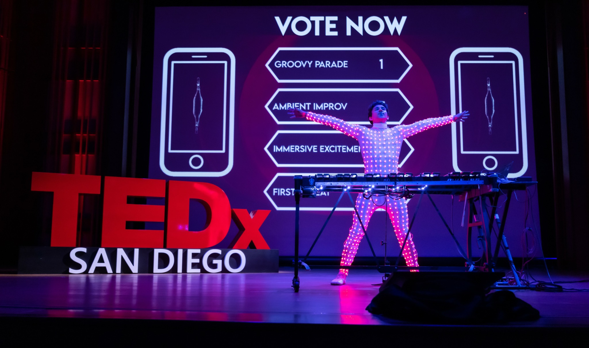 Person on stage with electronic equipment, large red TEDx sign, and a screen displaying voting options, in a dark auditorium.