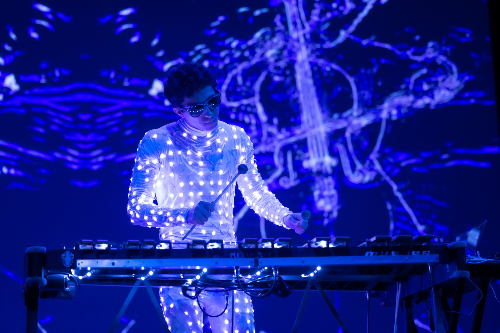 Person wearing glasses interacts with a digital music instrument in a dark environment with blue light projections.