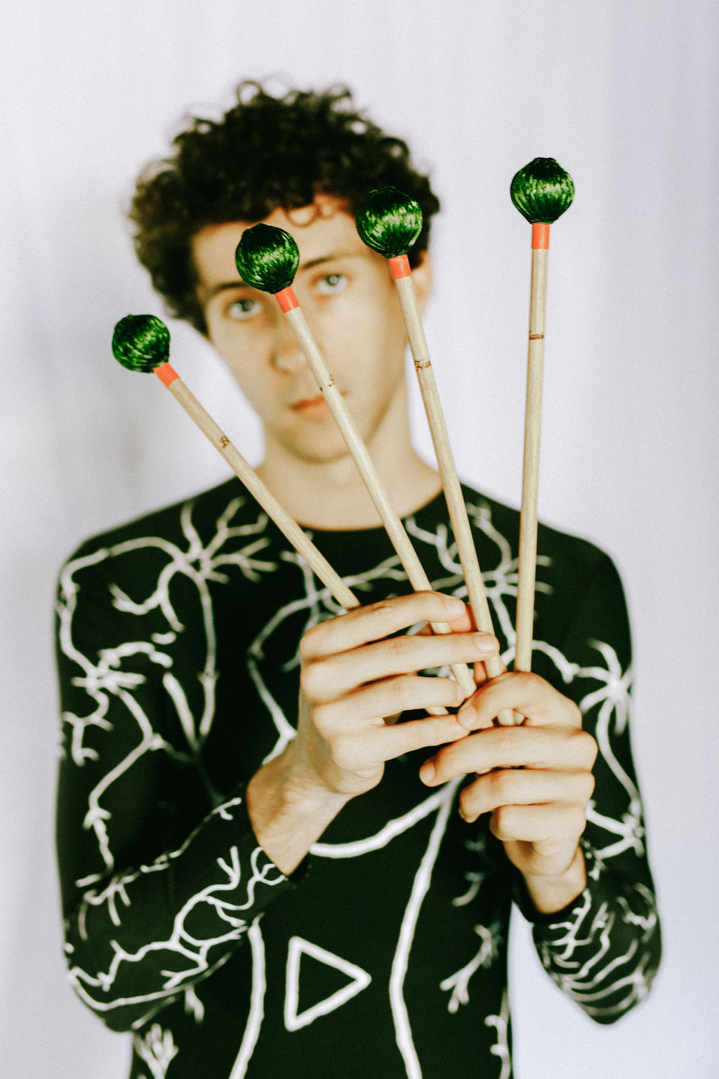 Person with curly hair holding four matchsticks with green balls on top, wearing a black shirt with white abstract patterns.