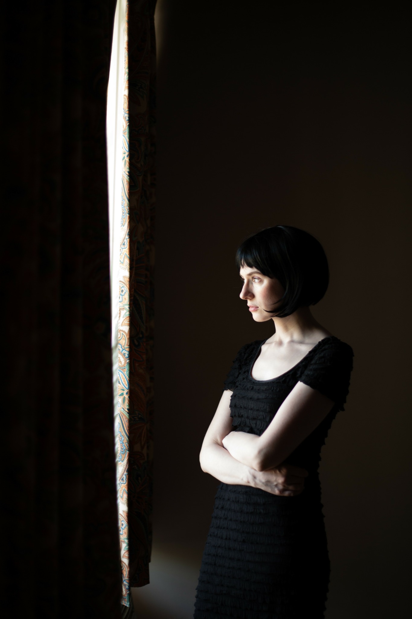 Woman with short dark hair stands near a window with a curtain, arms crossed, looking outside.