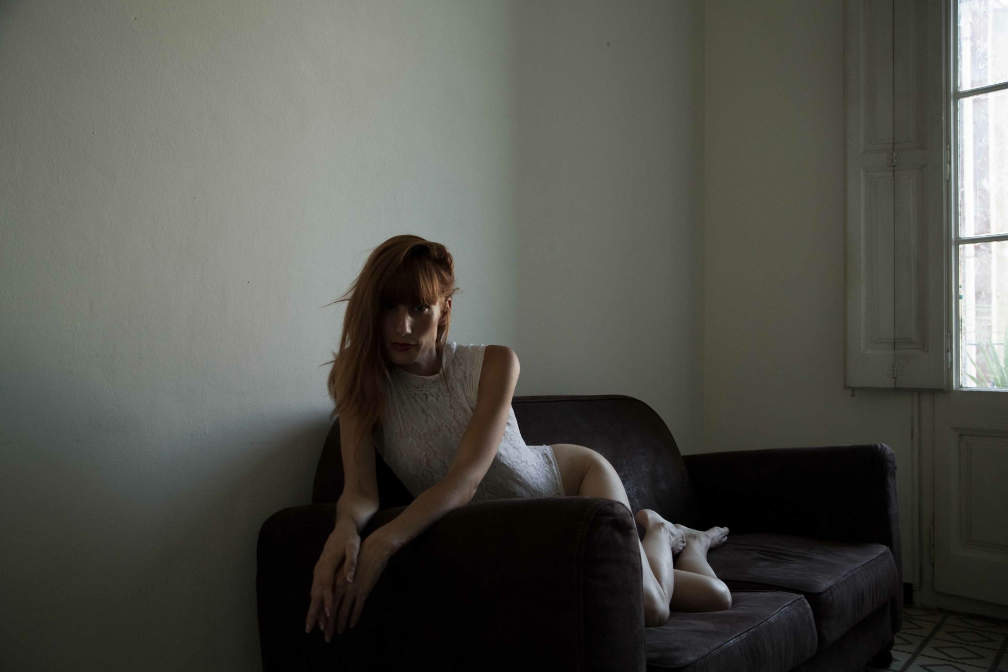 A woman with long hair sits on a dark sofa in a room with light-colored walls and a window with blinds.