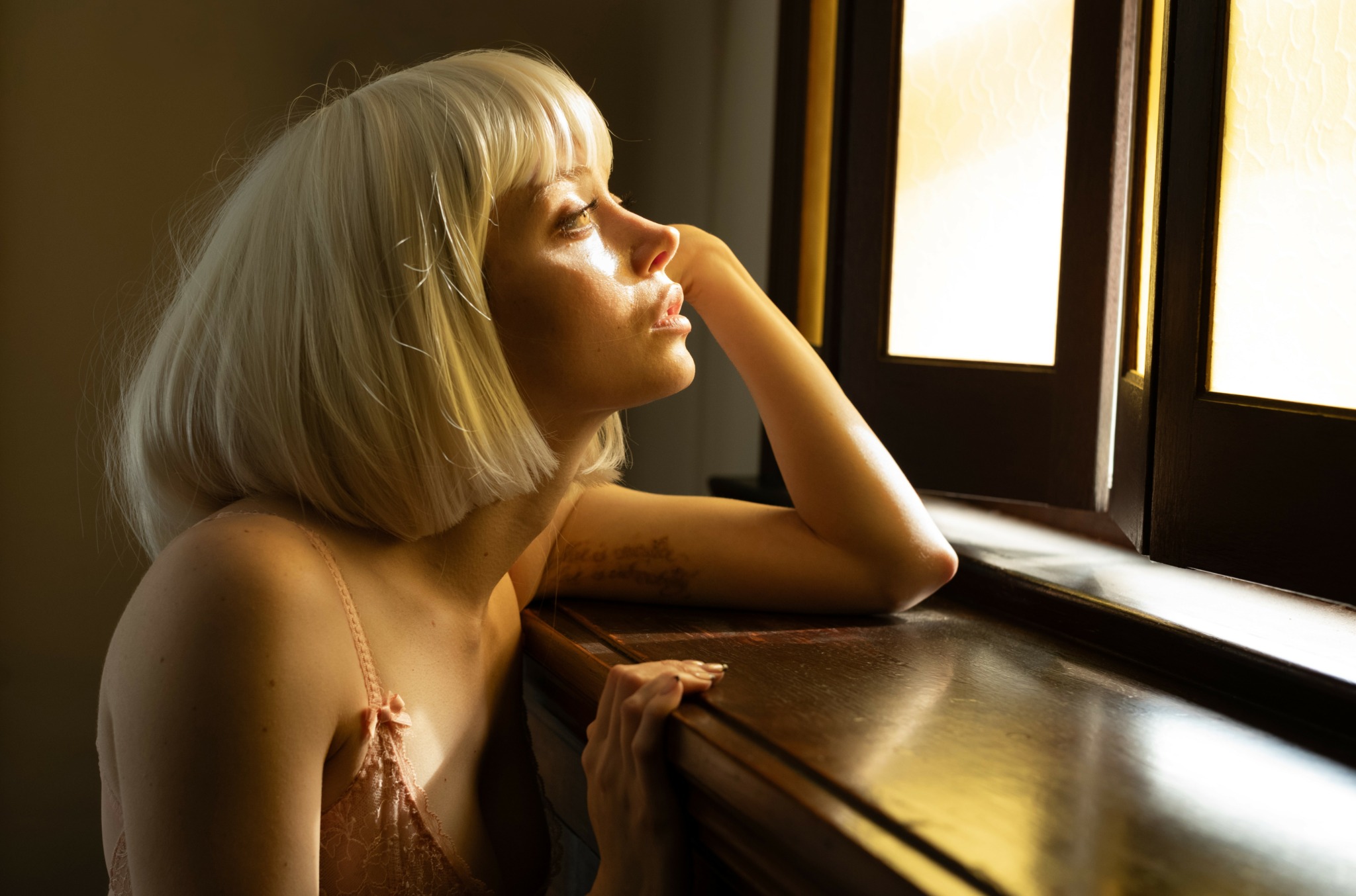 Woman with short blonde hair looking out a window, sunlight illuminating her face, resting her head on her hand.