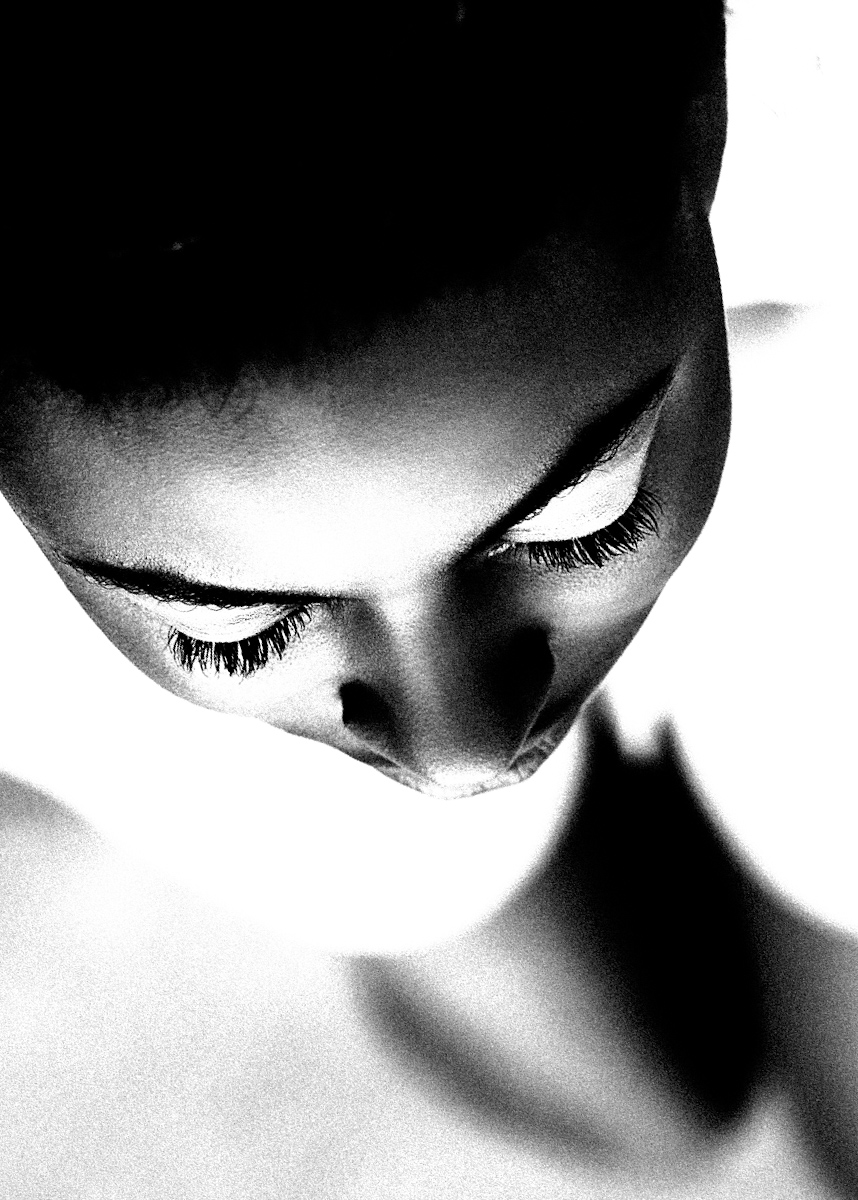 Close-up of a person's face looking downward, with long eyelashes and dark hair, in black and white.