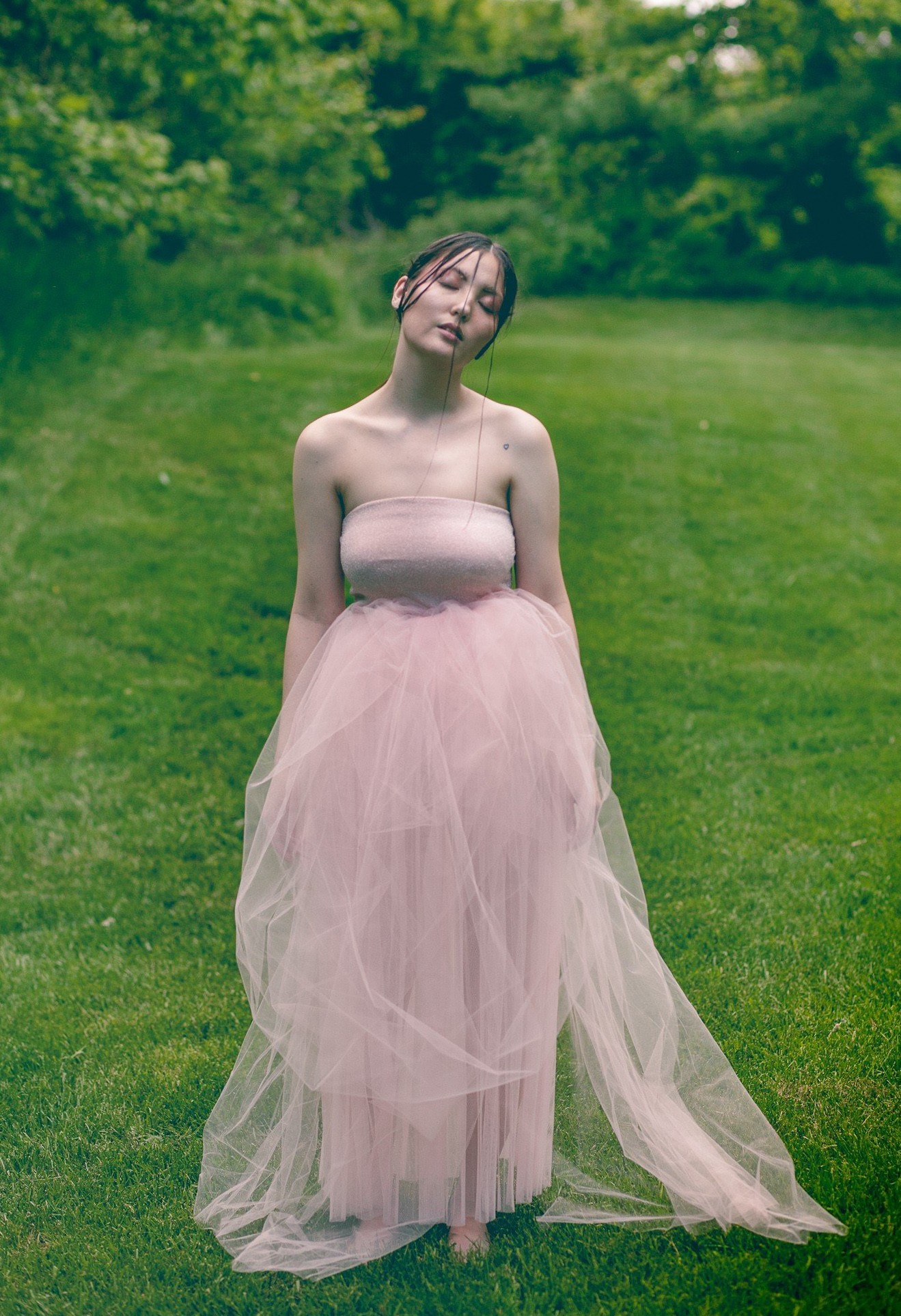 Young woman in a strapless pink dress standing on grass with trees in background.