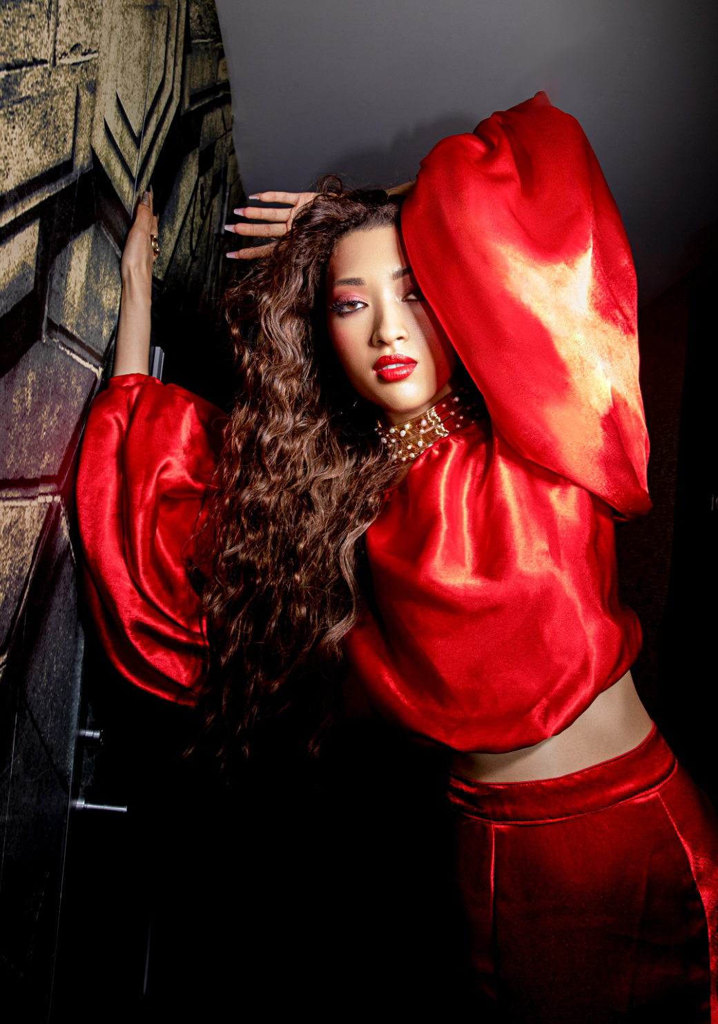 Woman with long curly hair wearing a red satin top and skirt, posing with one arm raised, against a dark background.