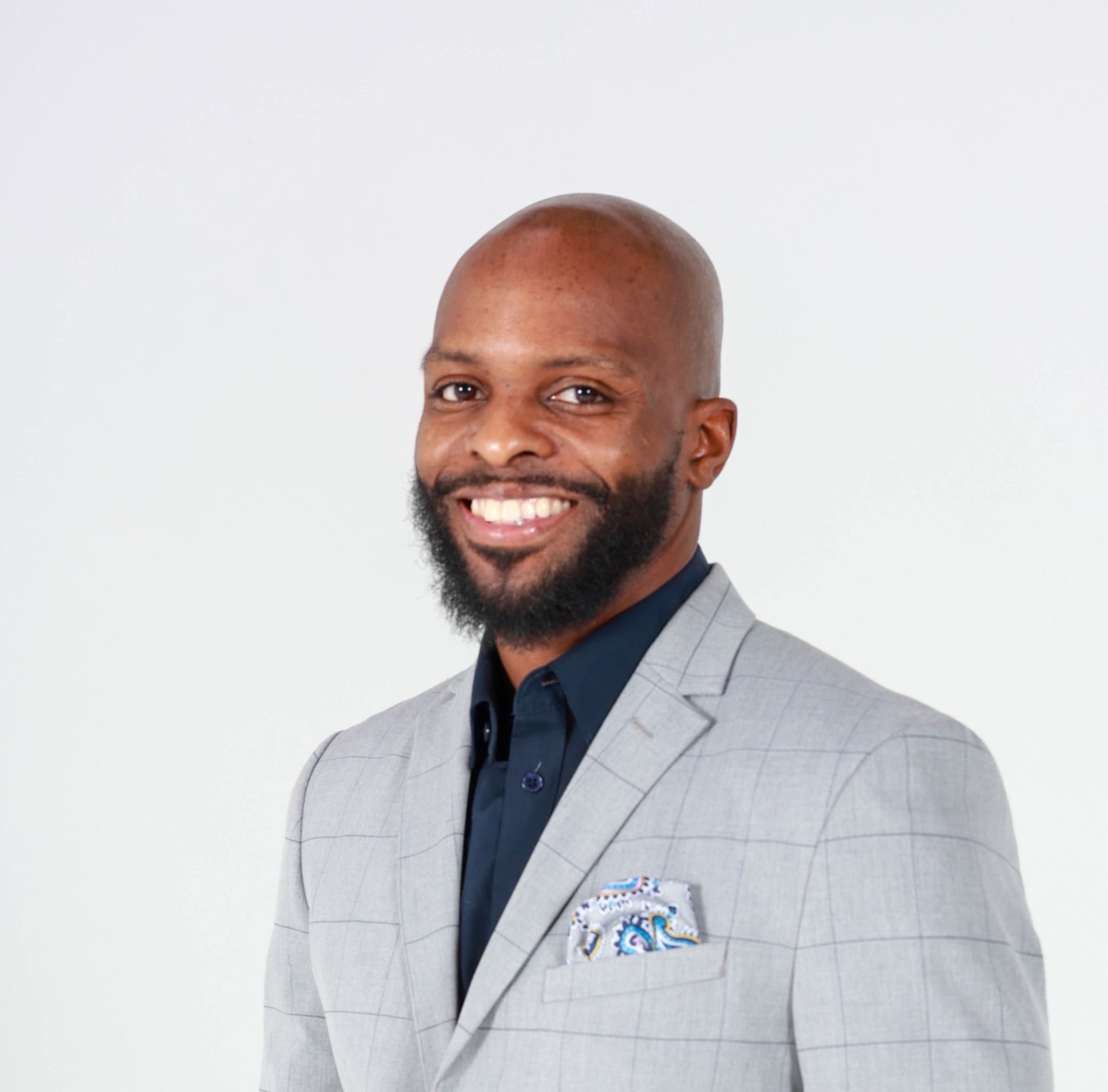 Man with a beard in a light gray suit and dark shirt, smiling, against a plain white background.