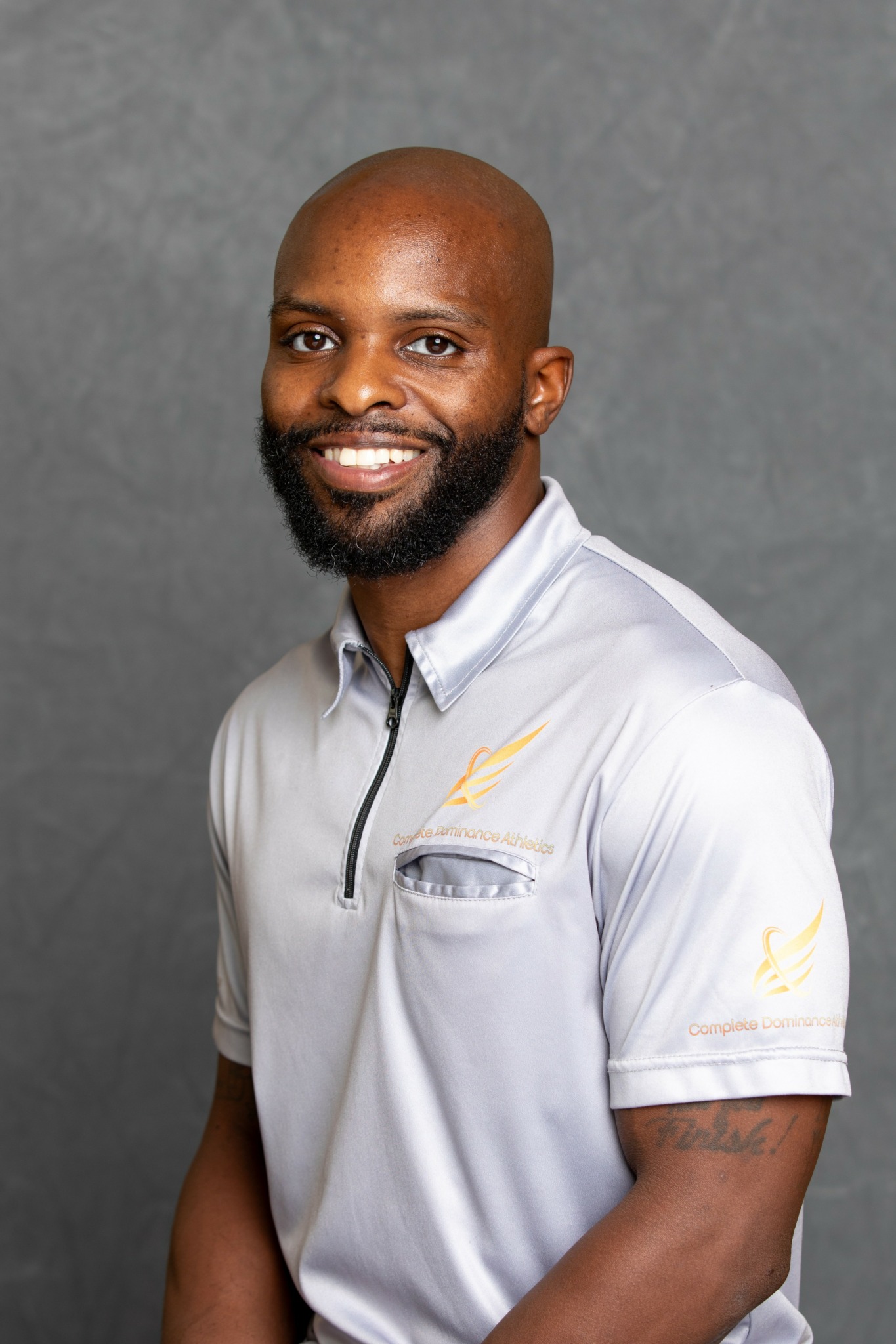 Smiling man with a beard and bald head wearing a light gray polo shirt against a gray background.