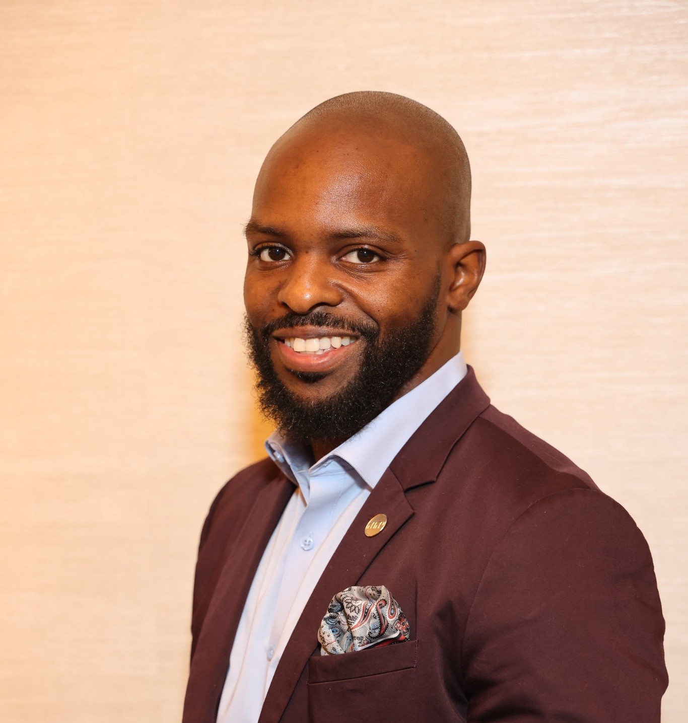 Man with a beard in a suit smiling, standing against a light background.