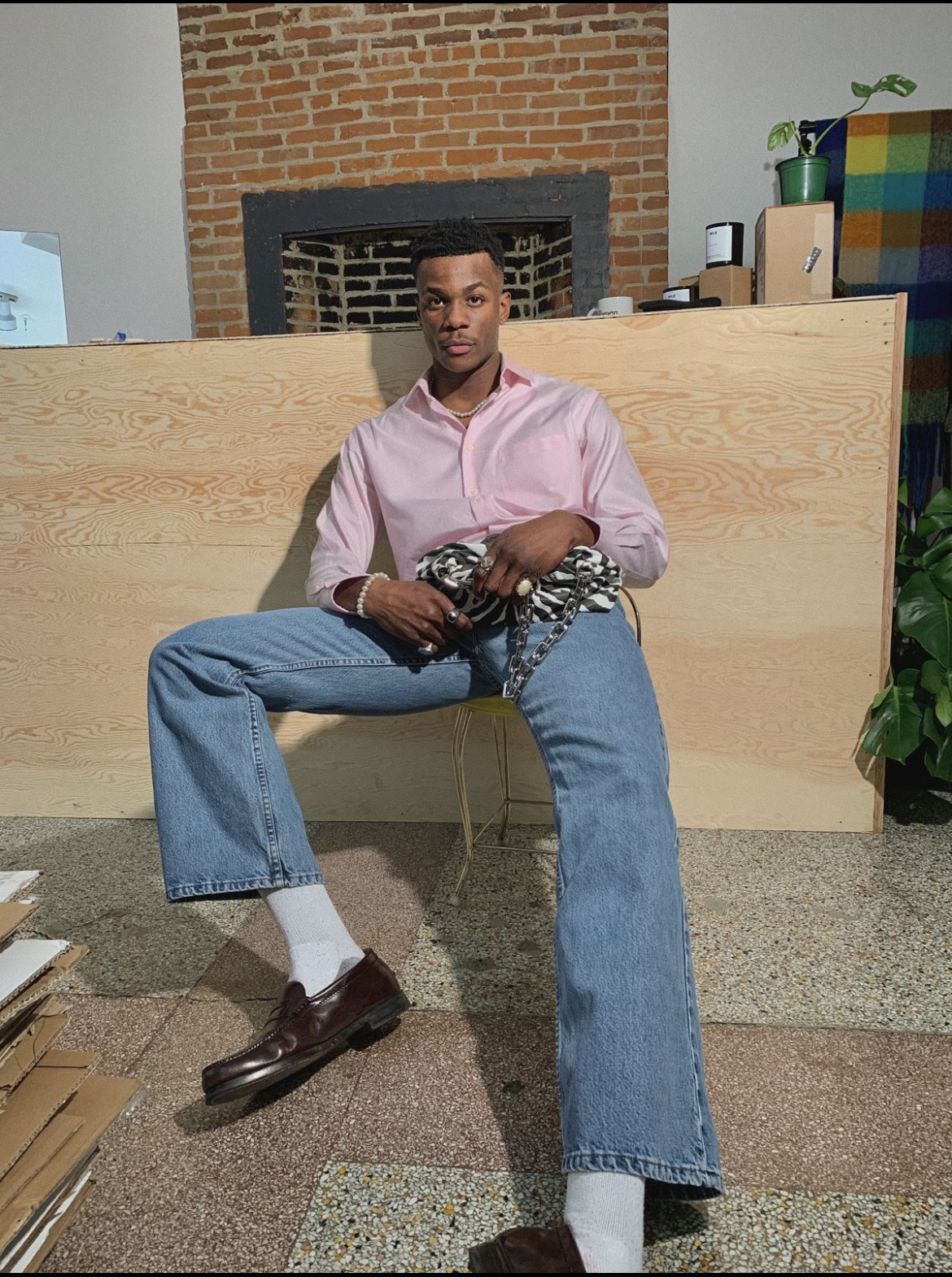 Person sitting on a chair in front of a brick fireplace, wearing a pink shirt, blue jeans, and brown shoes.