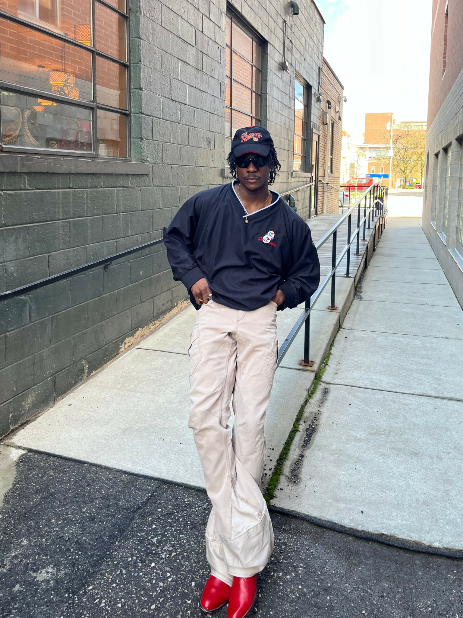 Person walking on sidewalk wearing black jacket, beige pants, red shoes, sunglasses, and a hat, with buildings in background.