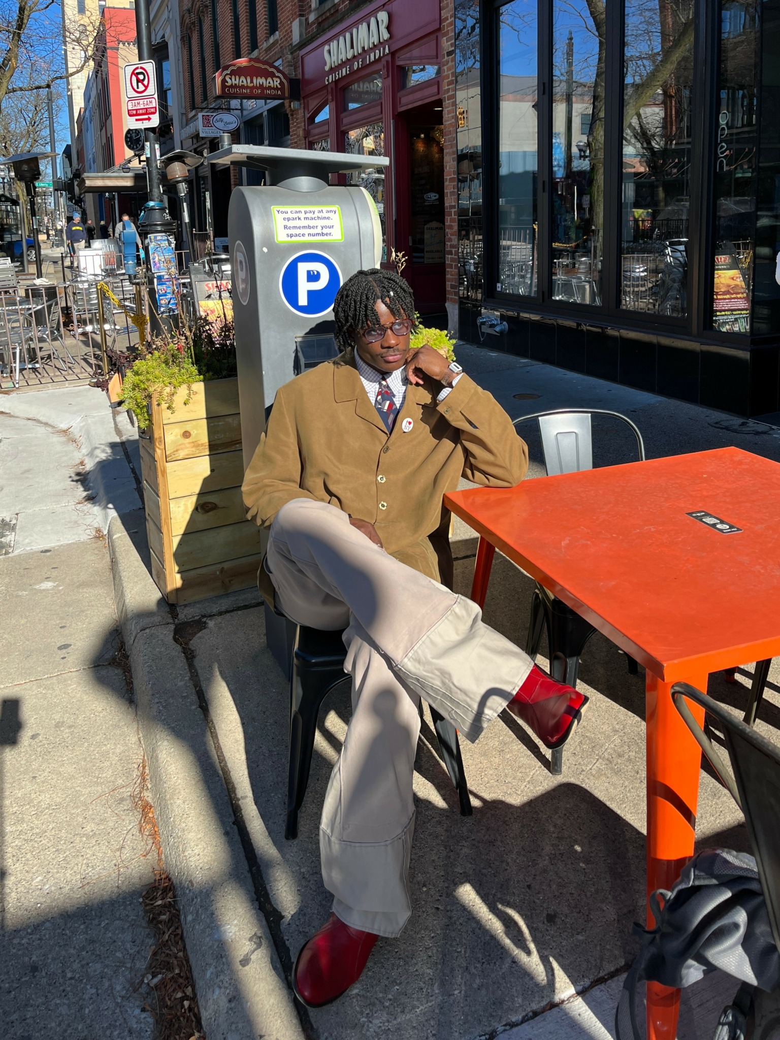 Person sitting at an outdoor table, eating ice cream, wearing a brown coat, beige pants, and red shoes, with a parking sign behind.