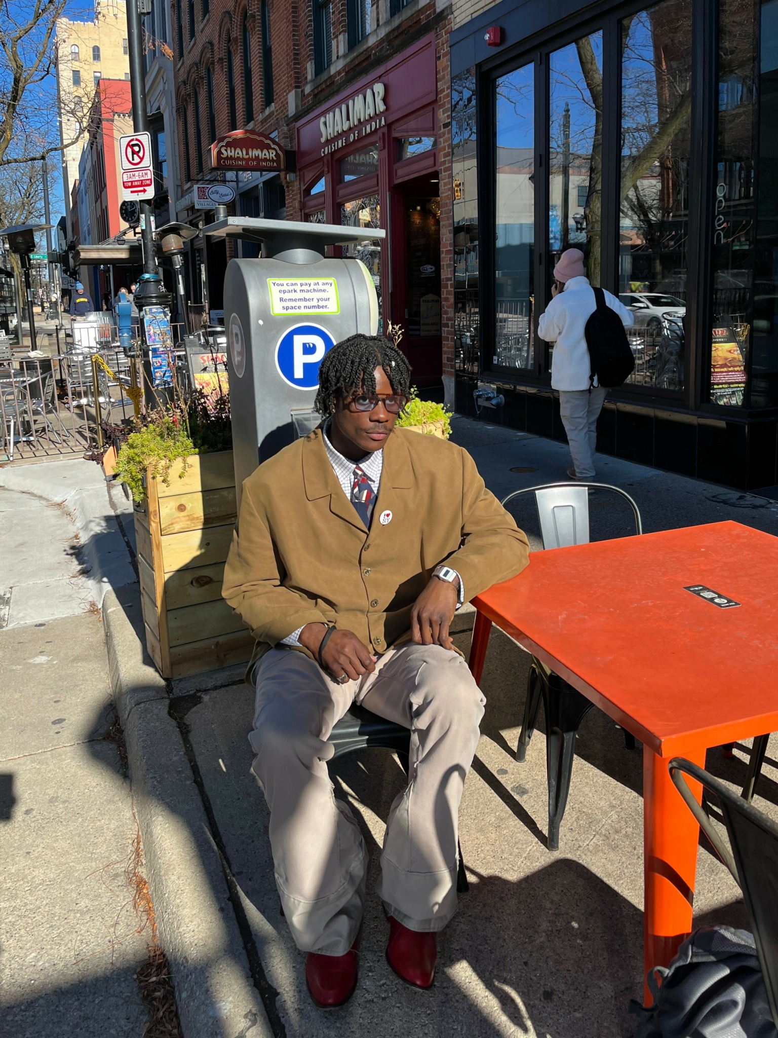 Person with glasses and dark hair sitting at an outdoor table on a city sidewalk, wearing a brown coat and red shoes.