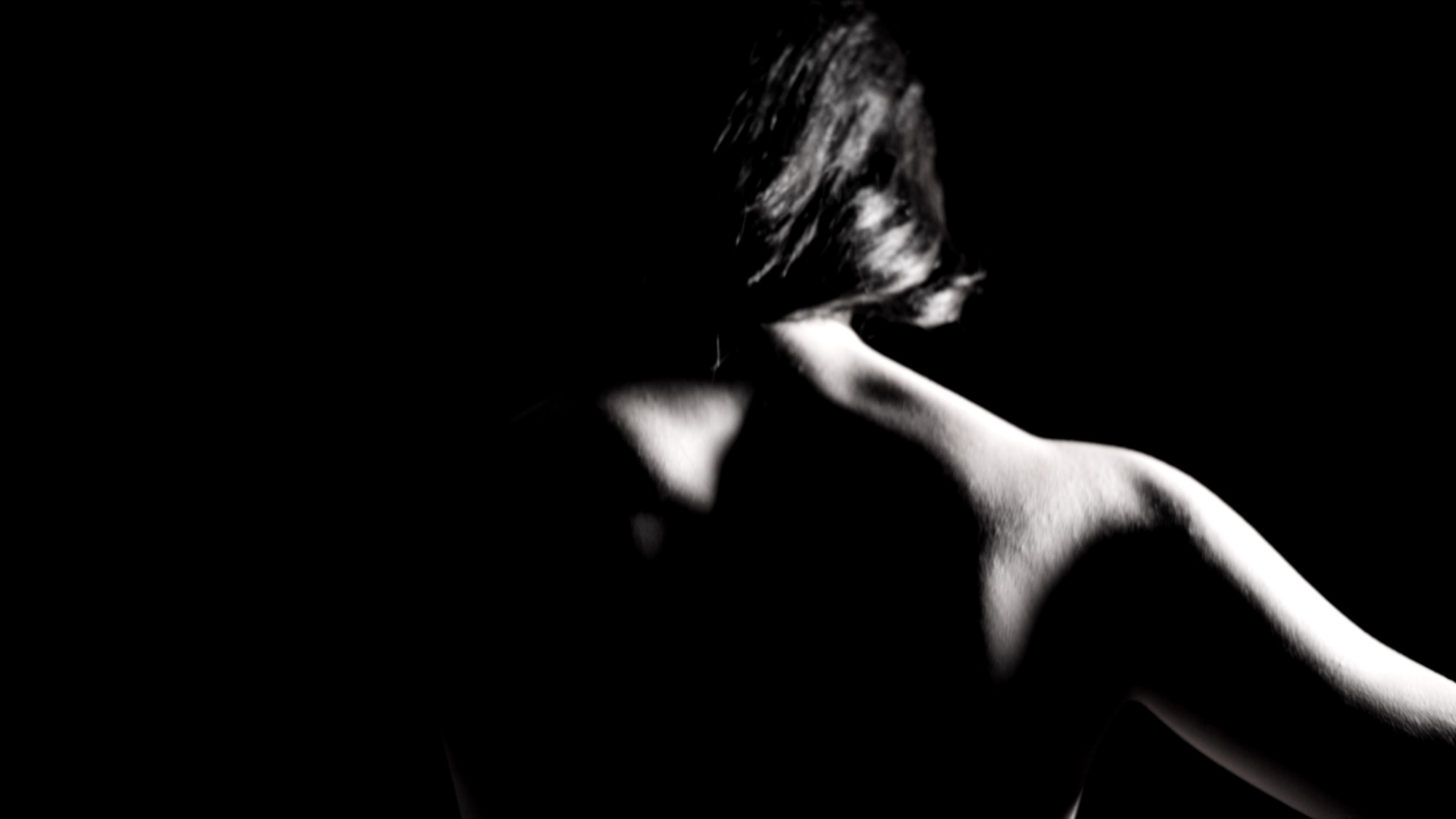 Back view of a person with short hair, illuminated shoulder and upper back, dark background.