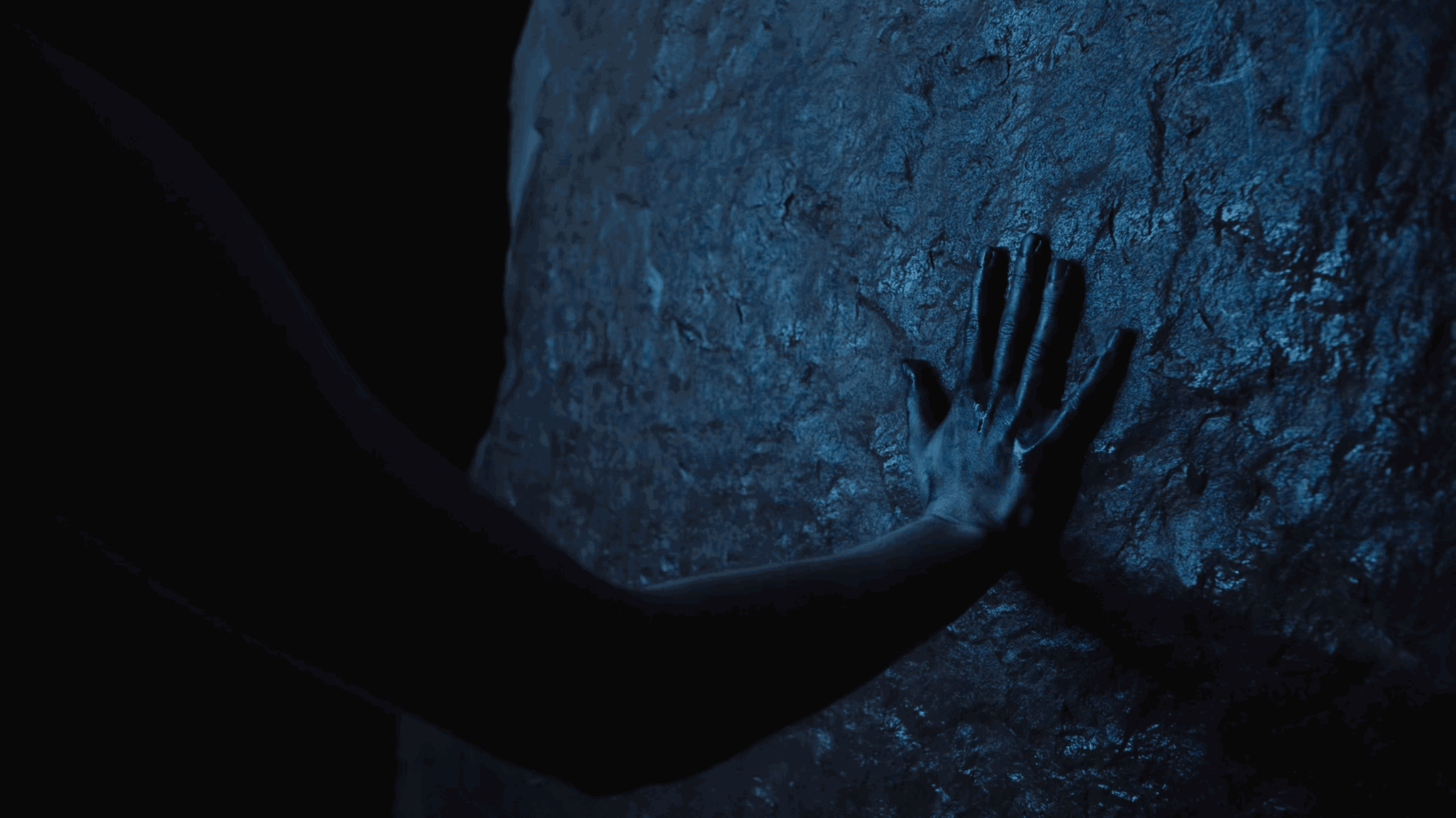 Person's hand touching a textured rock surface in low light, with part of their arm visible.