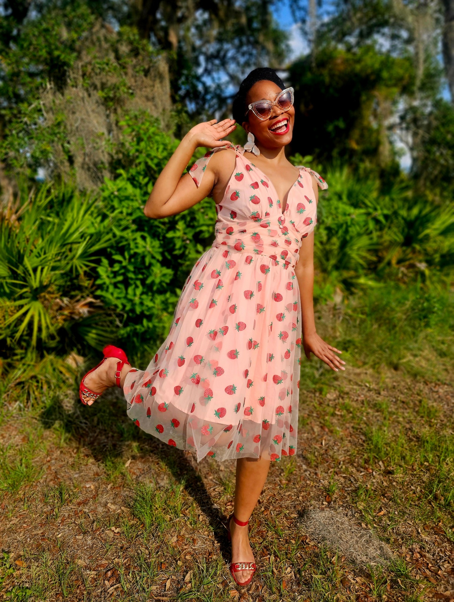 Woman in a pink polka dot dress smiling outdoors, holding her sunglasses, standing on grass with trees in background.