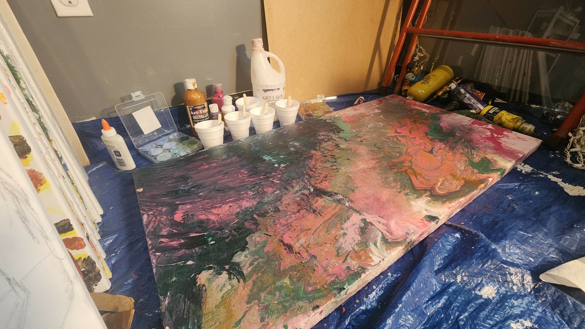 Large abstract painting with pink, green, and beige colors on a table covered with a blue tarp, surrounded by art supplies.