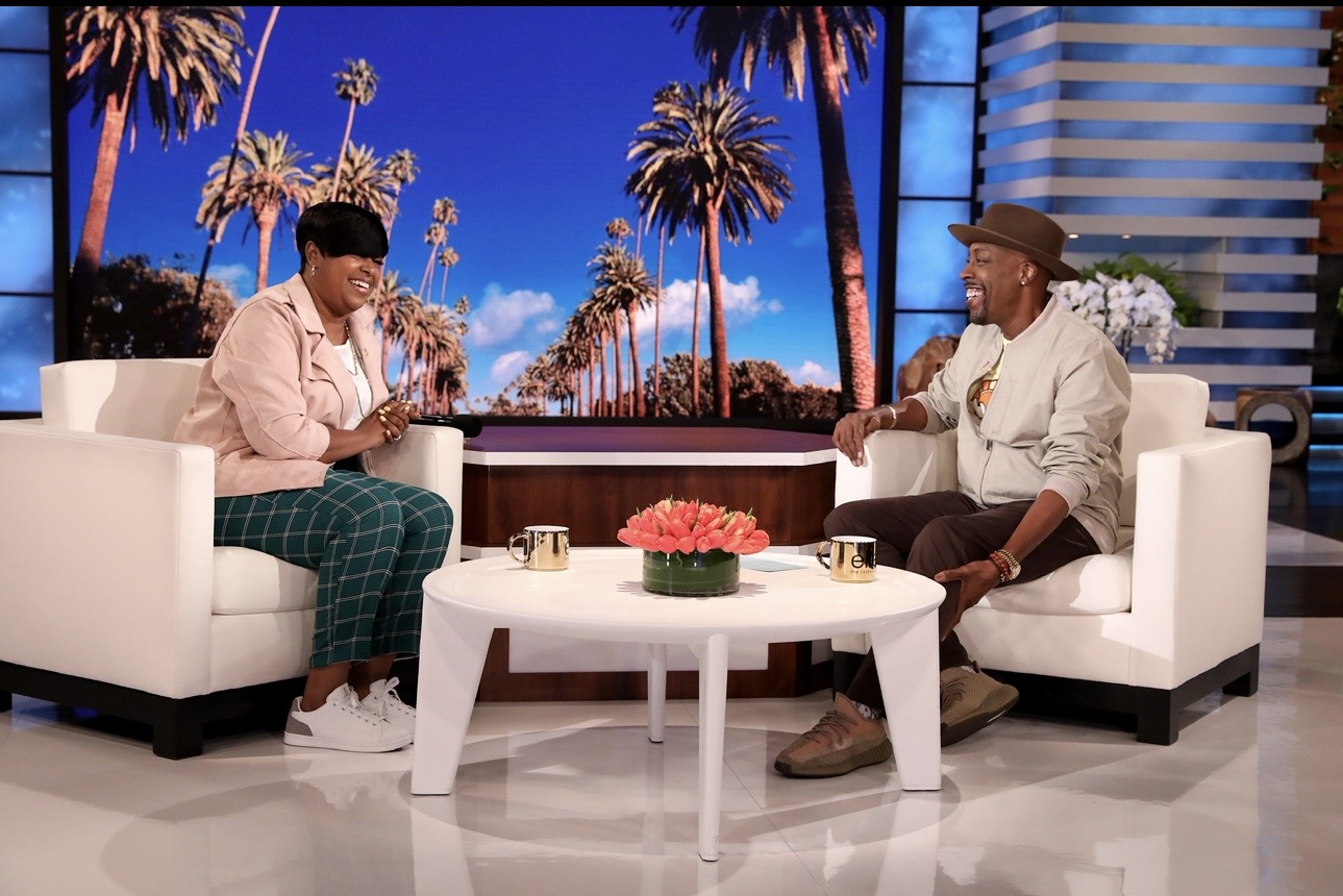 Two people sit on white armchairs in a studio with a backdrop of palm trees and blue sky, talking and drinking coffee.