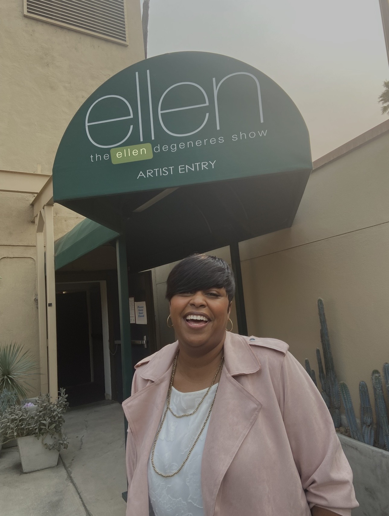 Smiling woman with short hair and a pink coat standing outside a building with a sign for Ellen Degeneres Show, artist entry.
