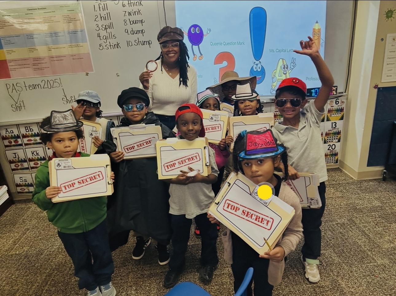 Group of children and an adult in a classroom holding boxes labeled 'Top Secret,' some wearing hats and sunglasses.