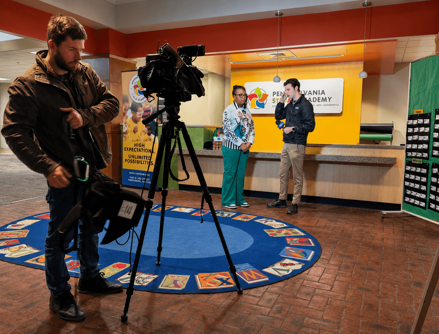 Two people stand in front of a reception desk with a large screen displaying a logo and text, while a cameraman films them.