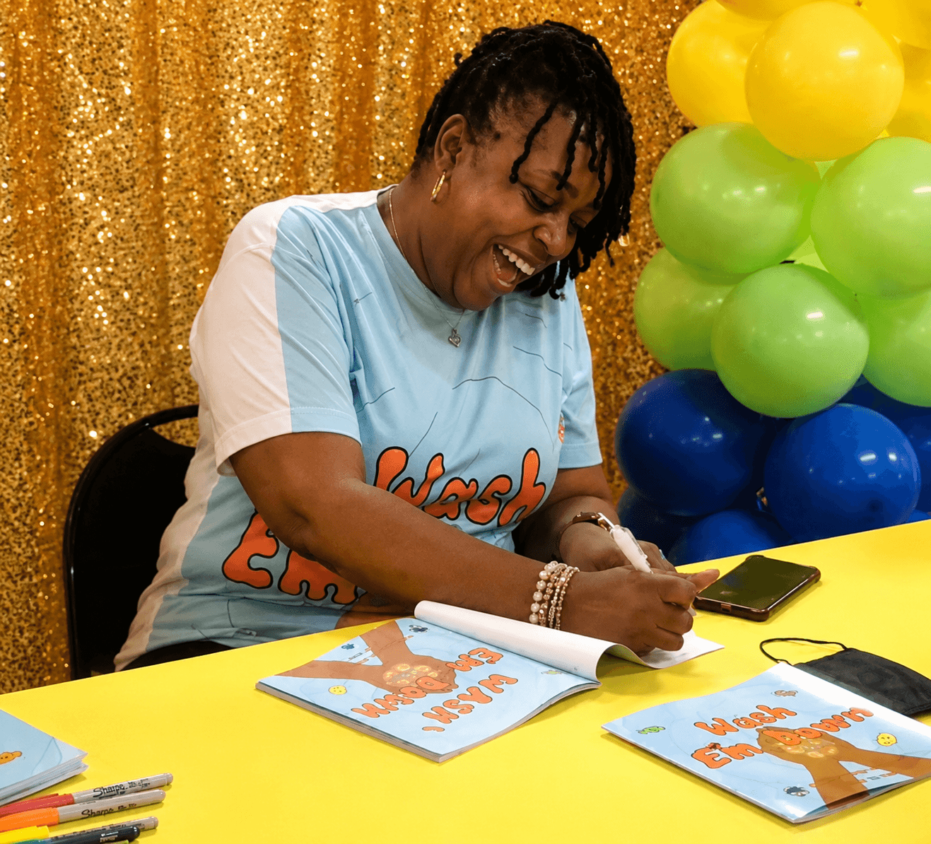 Person with short dreadlocks smiling, writing on a book at a table with colorful balloons in background.