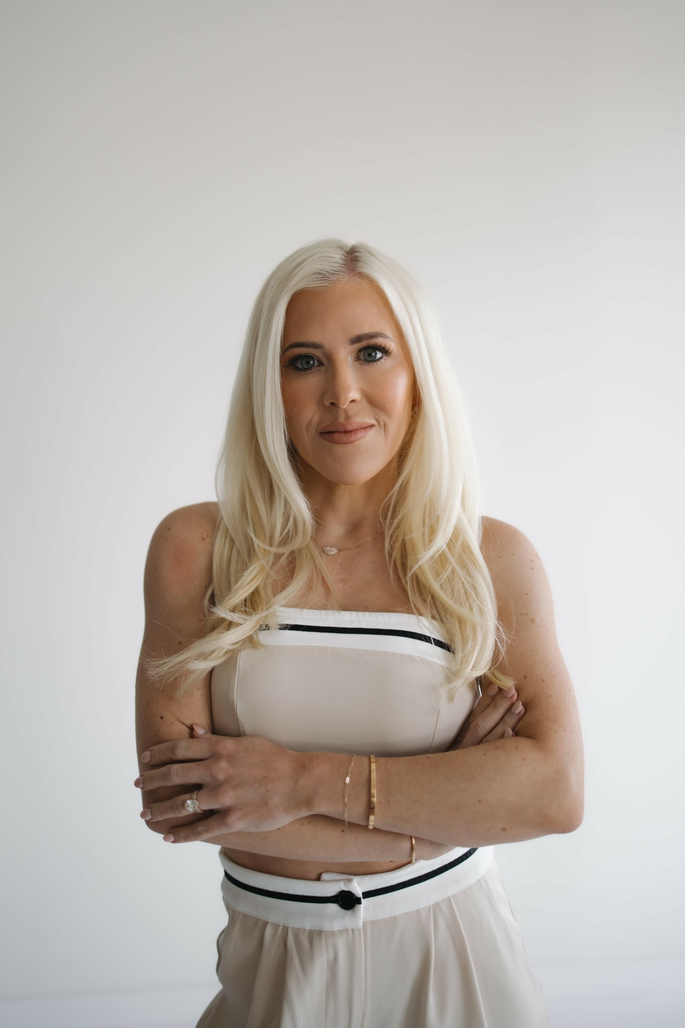 Woman with long blonde hair wearing a strapless top and light-colored pants, standing with arms crossed against a plain background.